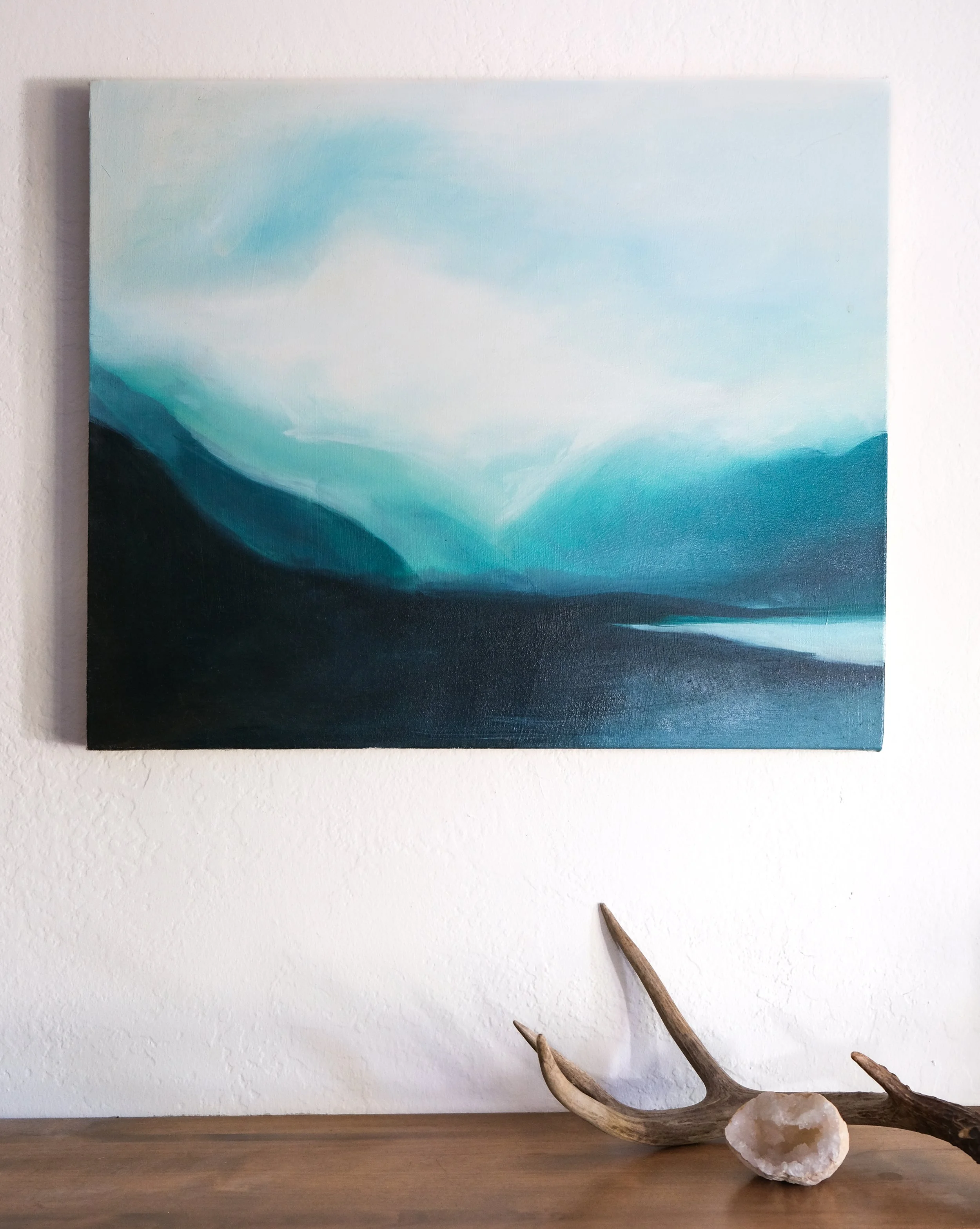 Decorative setup with abstract landscape painting on wall, antler with salt rock on wooden surface.