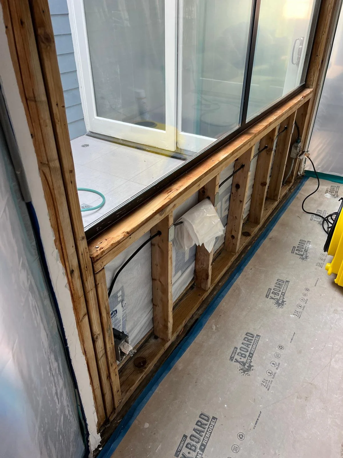 Construction of a framed wall with a glass sliding door, showing exposed wood studs and electrical wiring, in an indoor renovation area.