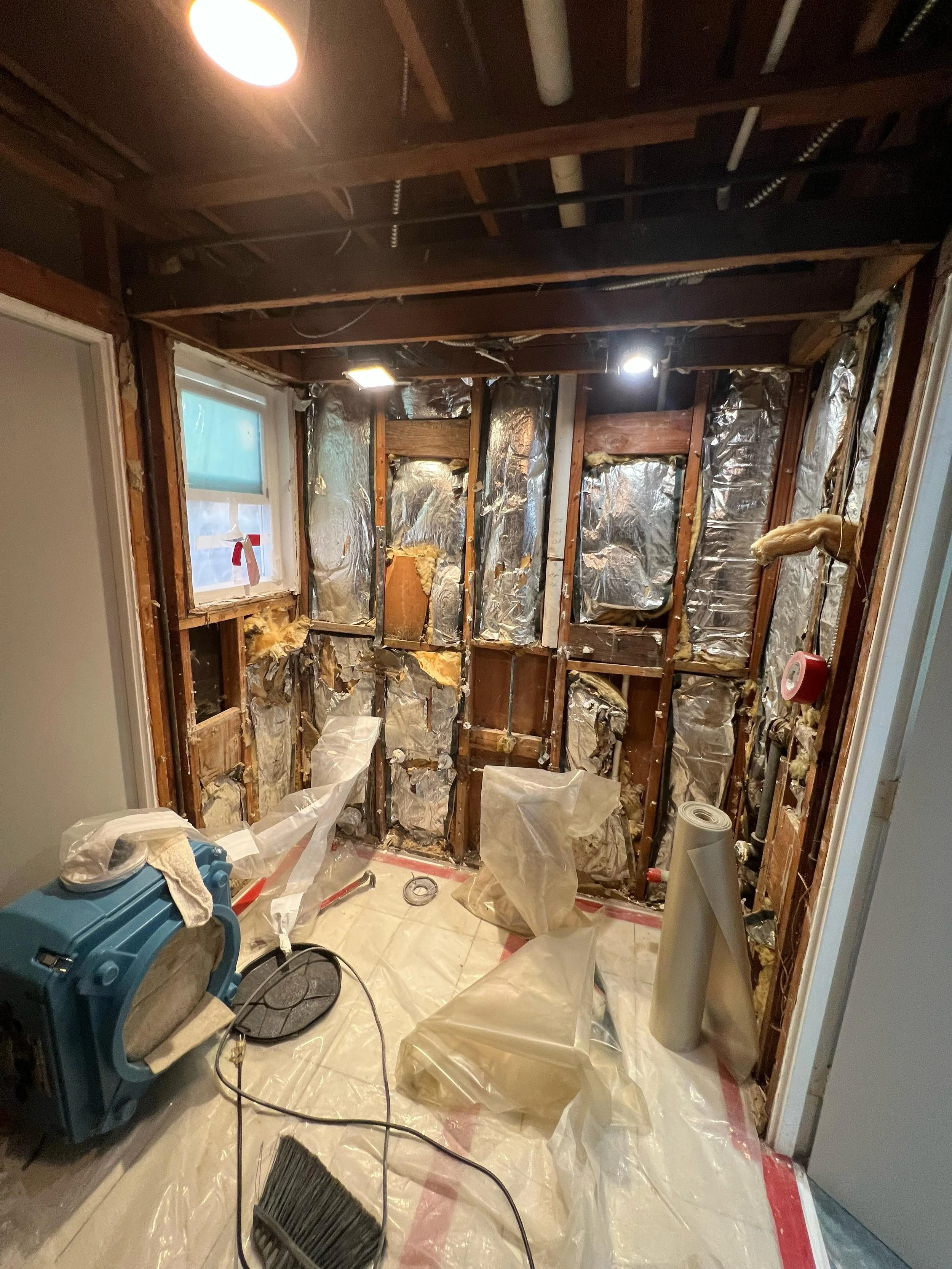 A room undergoing renovation with exposed wooden studs and insulation, trash bags, construction tools, and protective plastic covering the floor.