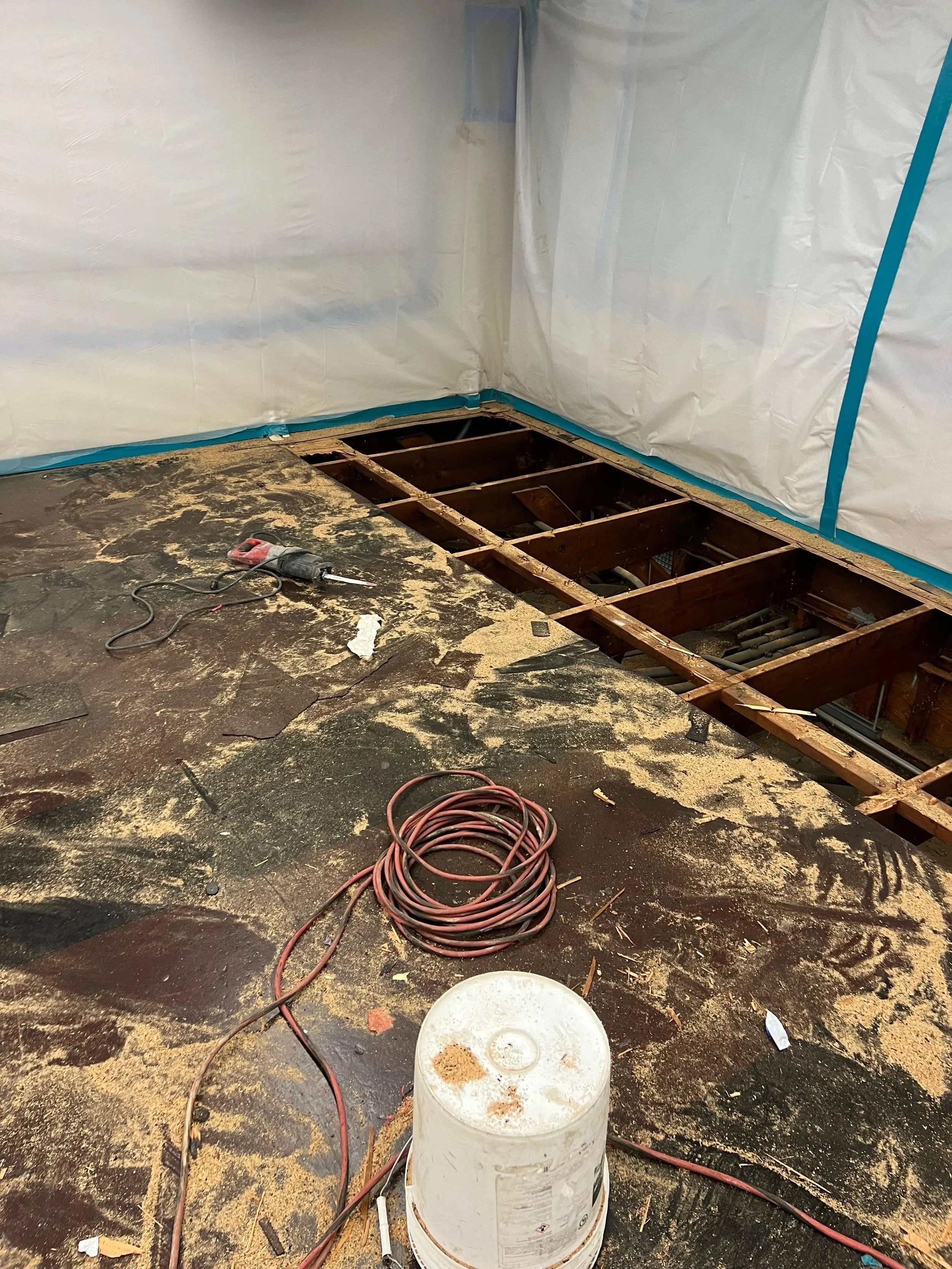 A construction or renovation site with a partially removed floor revealing wooden joists and plumbing underneath. The floor is dirty with debris, and there are tools and coiled electrical cords on the surface. In the background, white plastic sheetin