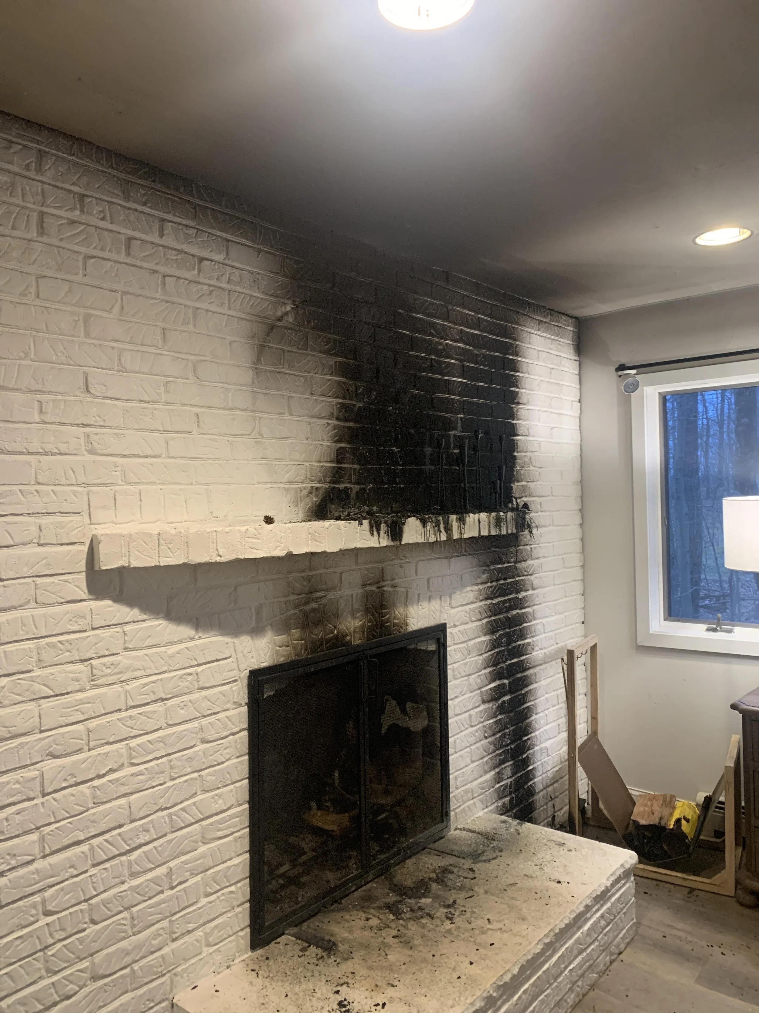 A white brick fireplace with blackened soot on the surface, with a firebox opening covered by a black mesh screen. The drywall above the fireplace appears clean, and a window with a white frame is visible to the right, showing a view of trees outside. The floor near the fireplace is cluttered with construction tools and materials.