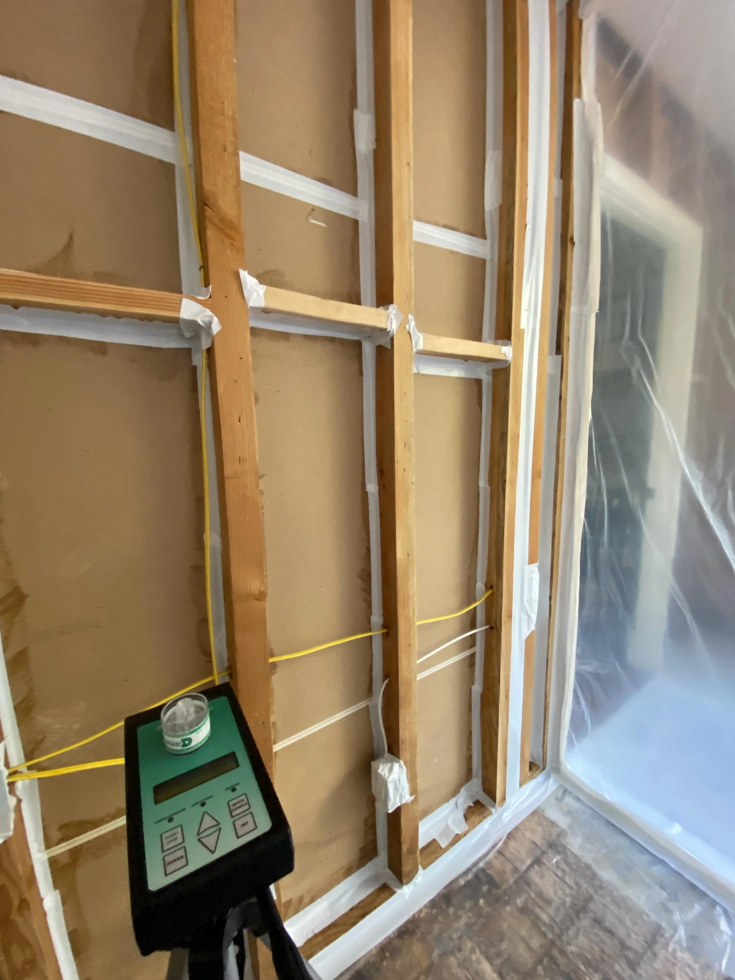 Interior view of a construction or renovation site with exposed wooden wall studs, some wiring, a digital device on a stand, and plastic sheeting covering part of the space.