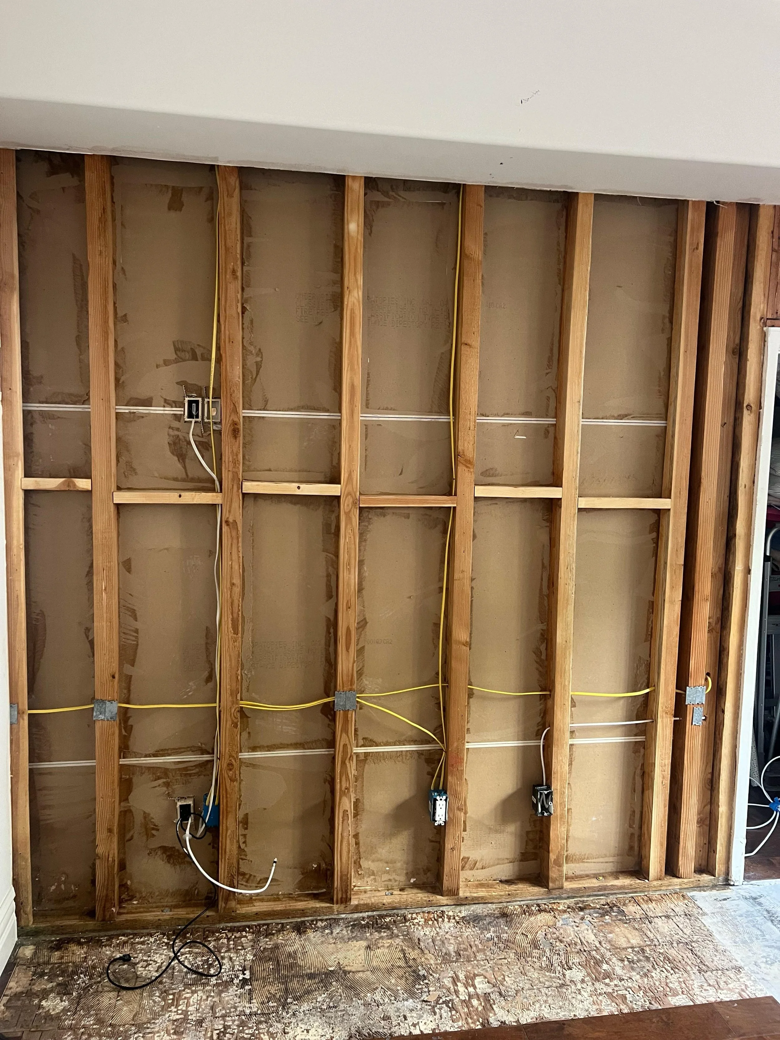 Open wall with exposed wooden studs, wiring, and electrical outlets, with a damaged floor in a room under renovation.