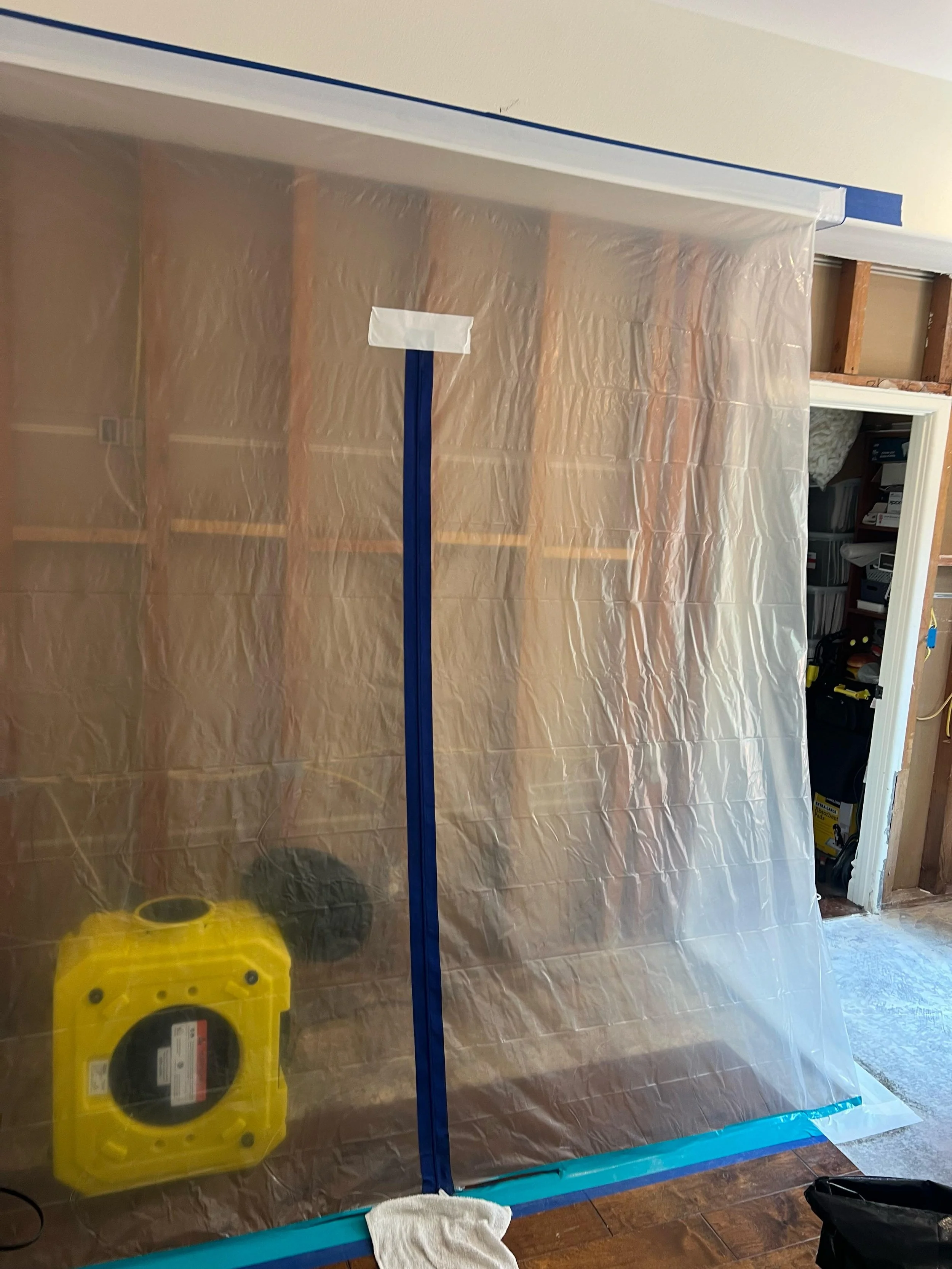 A plastic sheet hangs in front of a doorway, with blue painter's tape securing the top and bottom edges. Inside the room, there are construction tools and materials including a yellow portable heater and a white cloth on the floor. The room appears t