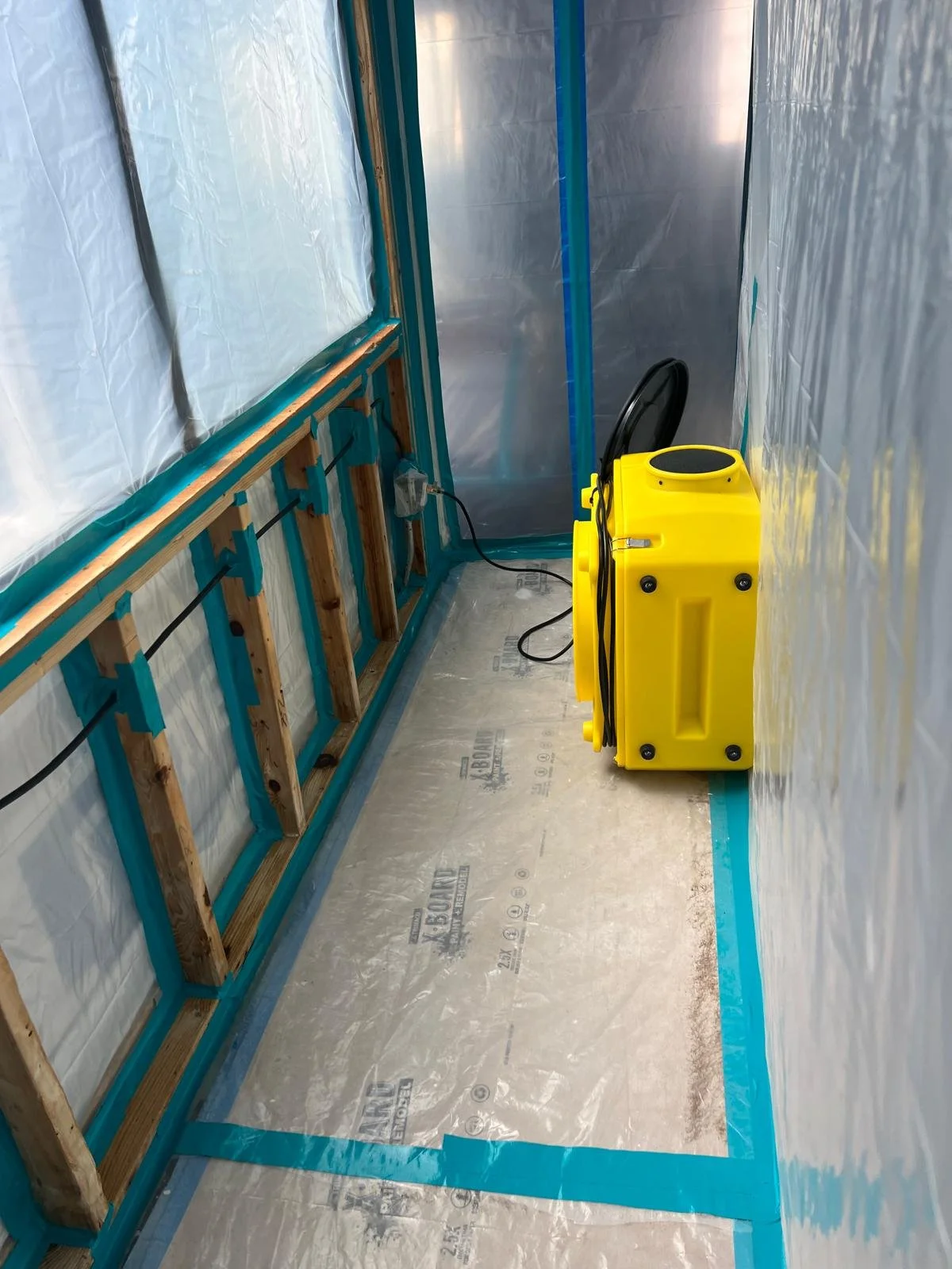 Interior of a small bathroom under construction with exposed wall studs, plastic sheeting on the walls, a yellow portable heater or dehumidifier, and an electrical outlet with a plugged-in cord.