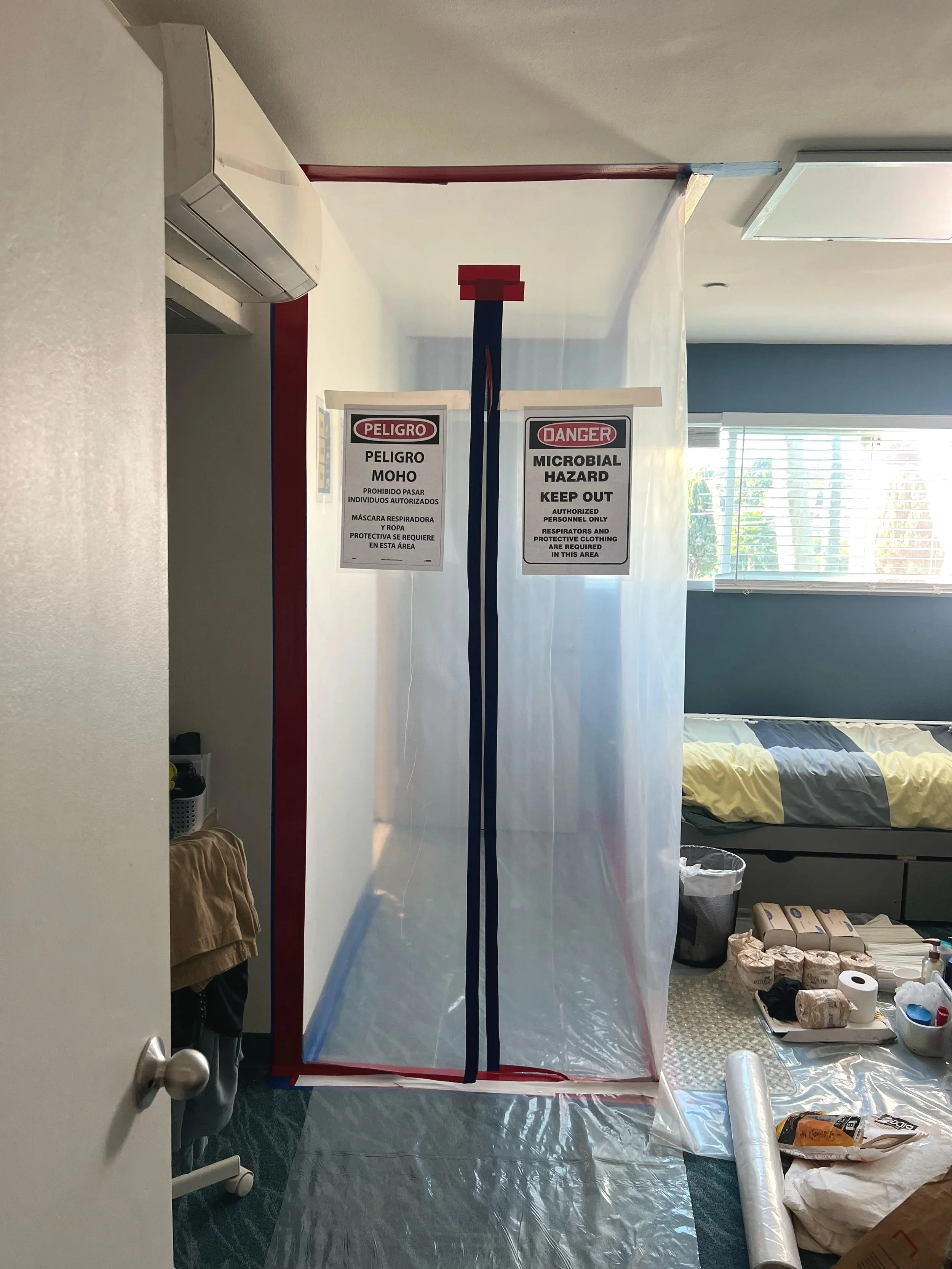 A transparent plastic sheet partitions a room, with warning signs that read 'Peligro Moho' and 'Danger Microbial Hazard Keep Out.' Inside, there are cleaning supplies, paper towels, and rolls of plastic wrap on a metal table, with a bed and window in the background.