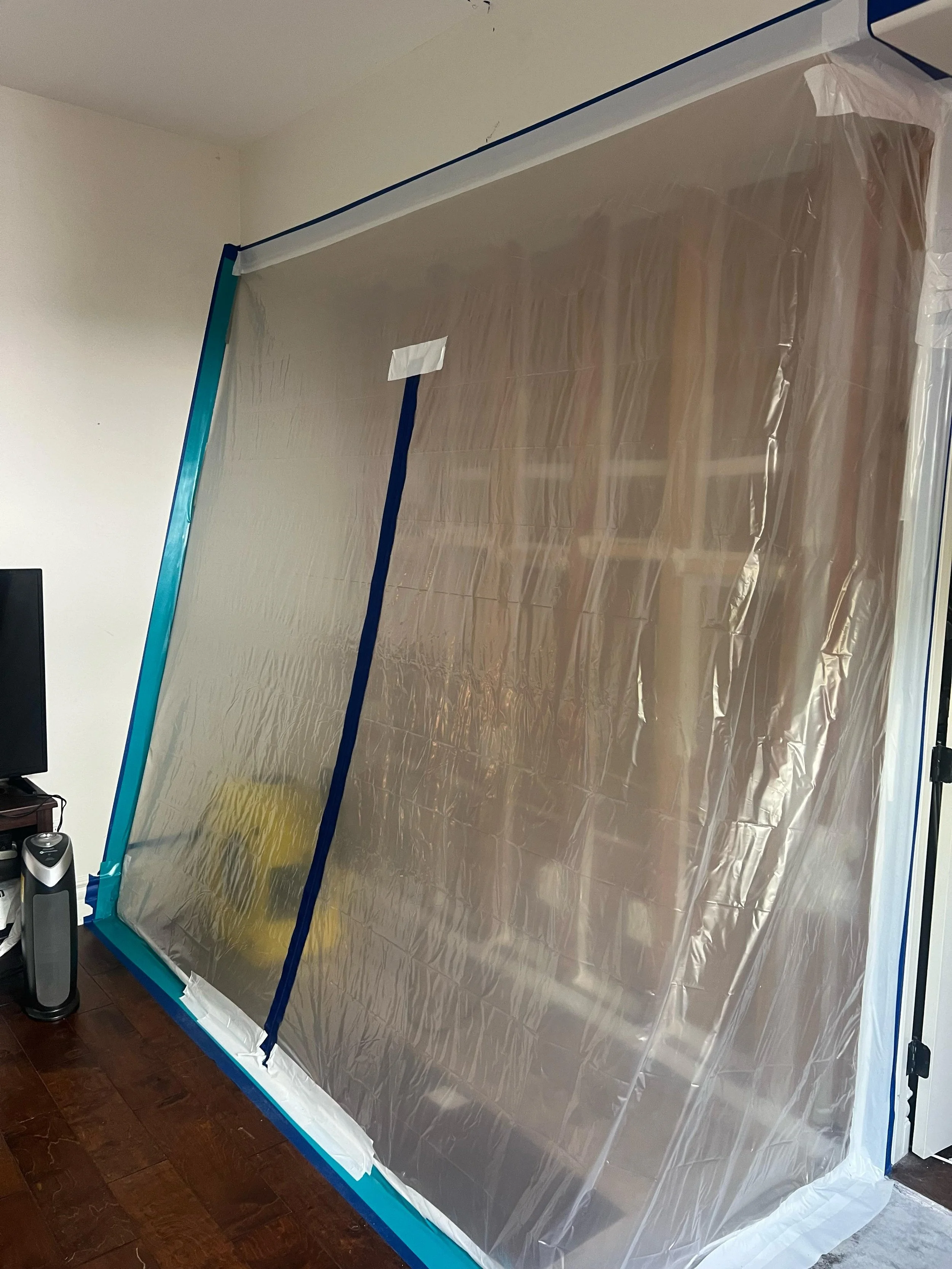 A plastic sheet hung to protect the wall and furniture during painting, with blue masking tape securing the edges. Part of a monitor and a tower are visible in the bottom left corner, on a dark wooden floor.