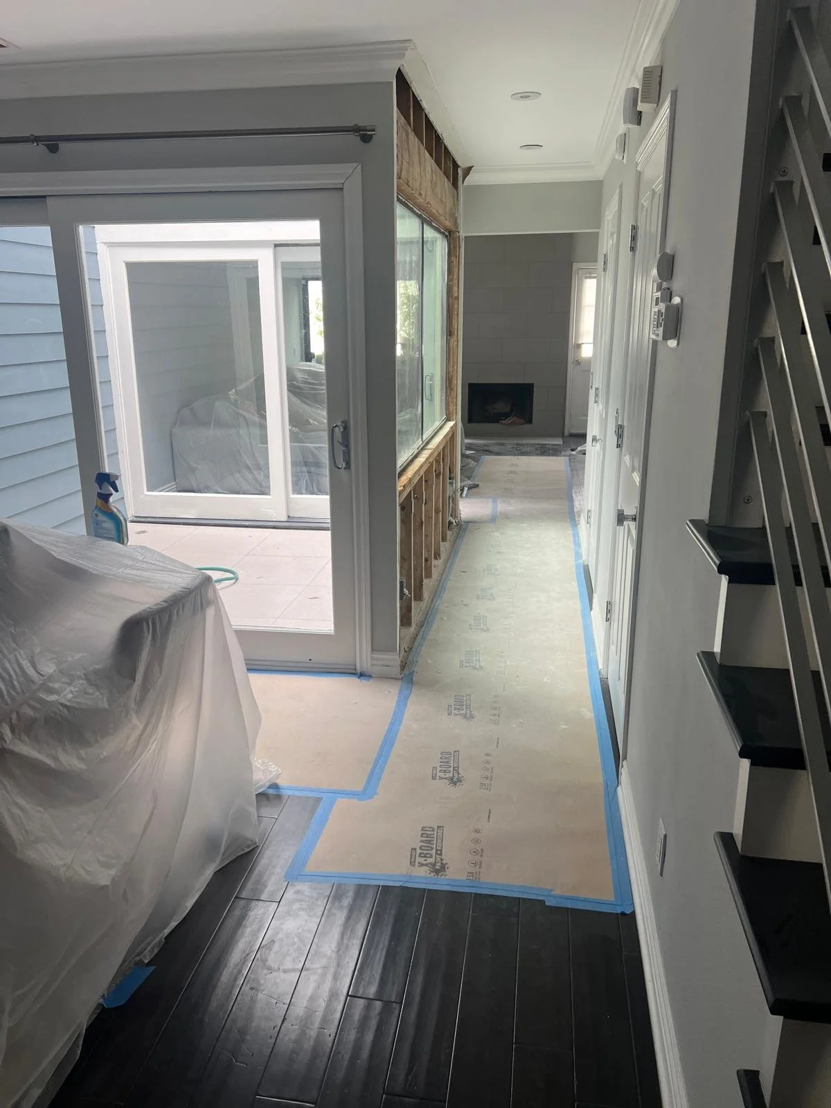 A room under renovation with protective floor covering, a sliding glass door leading to a patio, and an unfinished wall area with exposed studs.