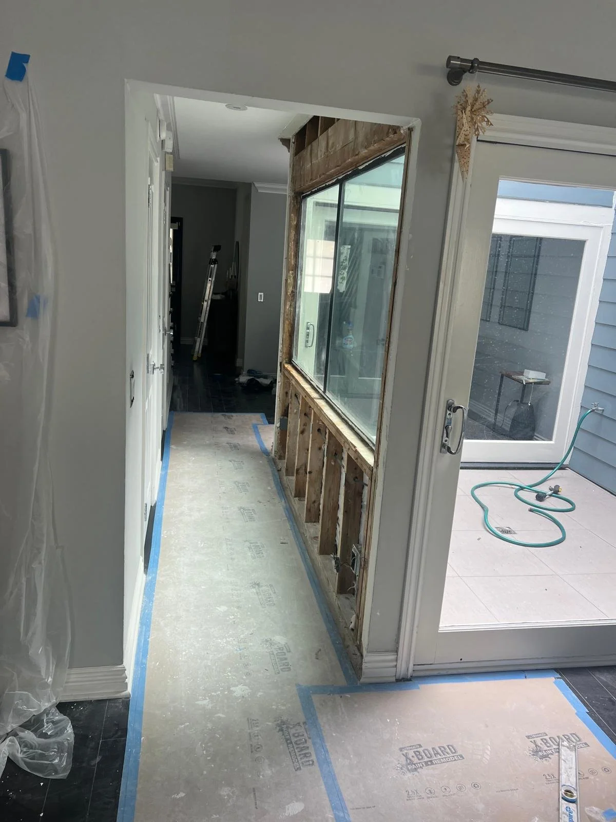 Open doorway showing a kitchen renovation with a partially removed wall, exposing wooden studs and a large window. A sliding glass door leads to a patio area with a garden hose, and construction tools and materials are visible throughout the space.