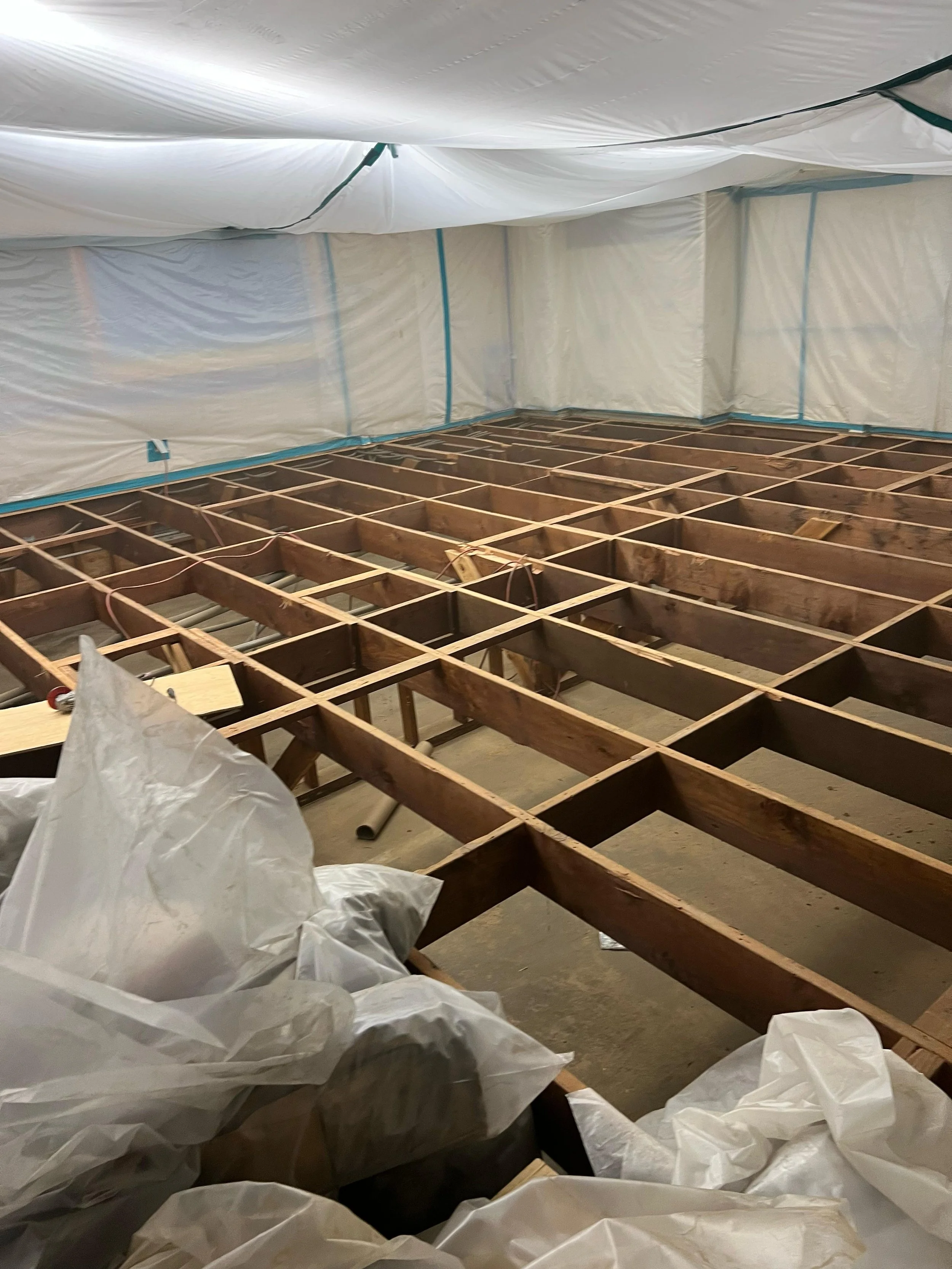 Construction site with wooden framework and white plastic sheeting walls and ceiling, with electrical wires and plastic bags on the floor.