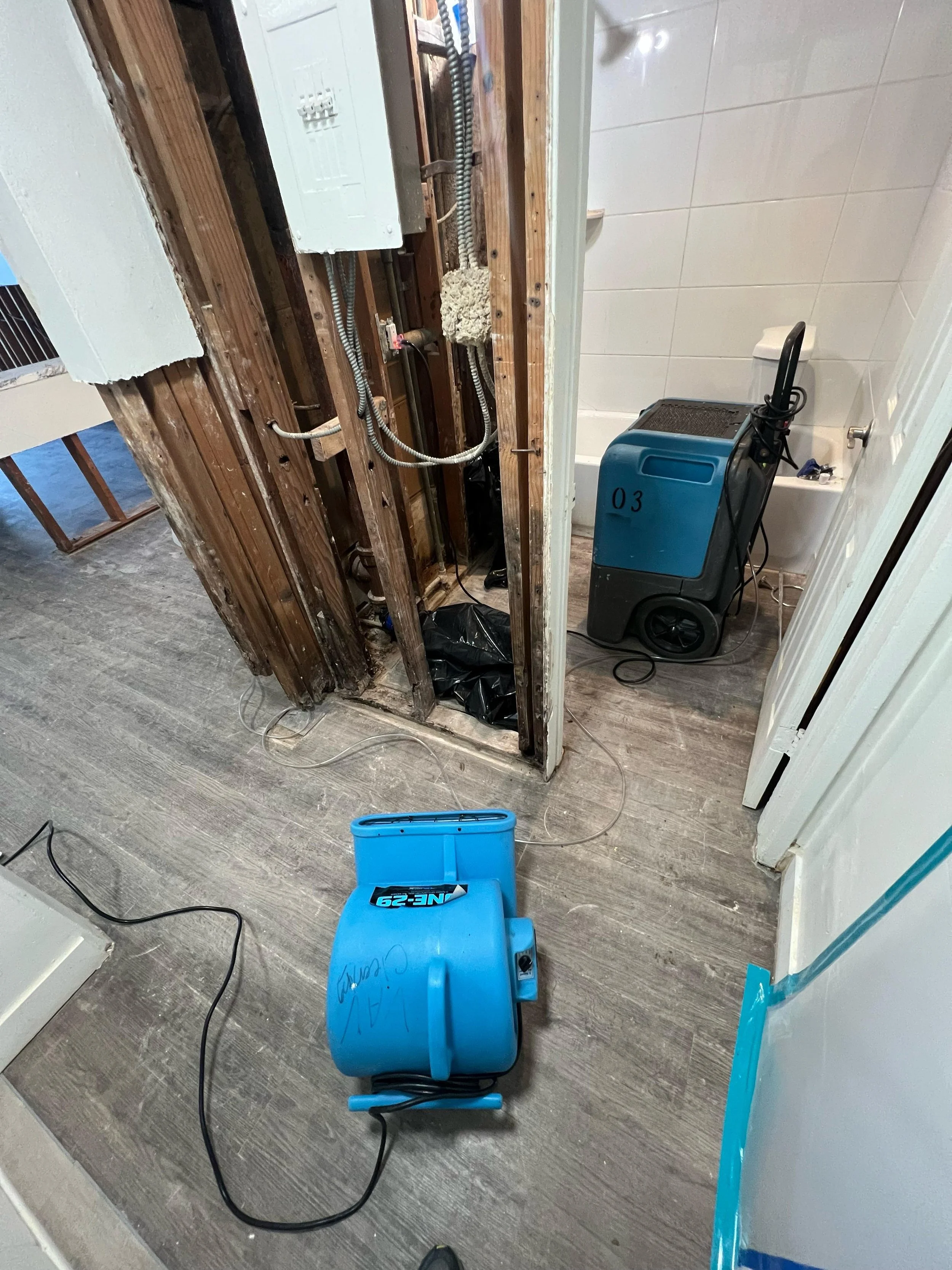 Bathroom undergoing renovation with exposed wooden wall studs, electrical wiring, and plumbing. A blue wet vacuum cleaner, labeled '62-29,' is on the floor, along with a blue toolbox, wires, and construction debris.