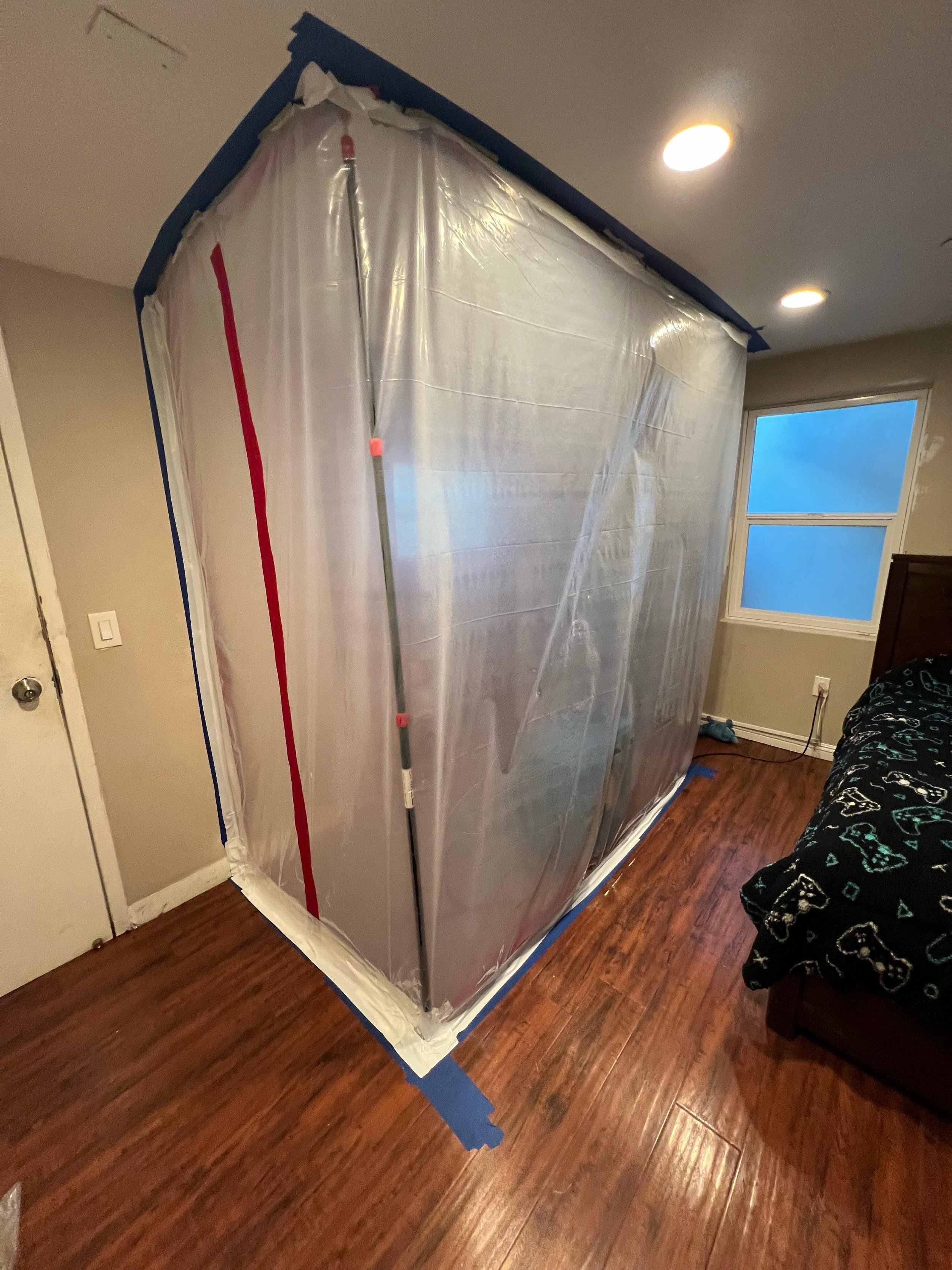A large cardboard box covered with clear plastic in a room with hardwood floors and a window, with a broom leaning against the box.