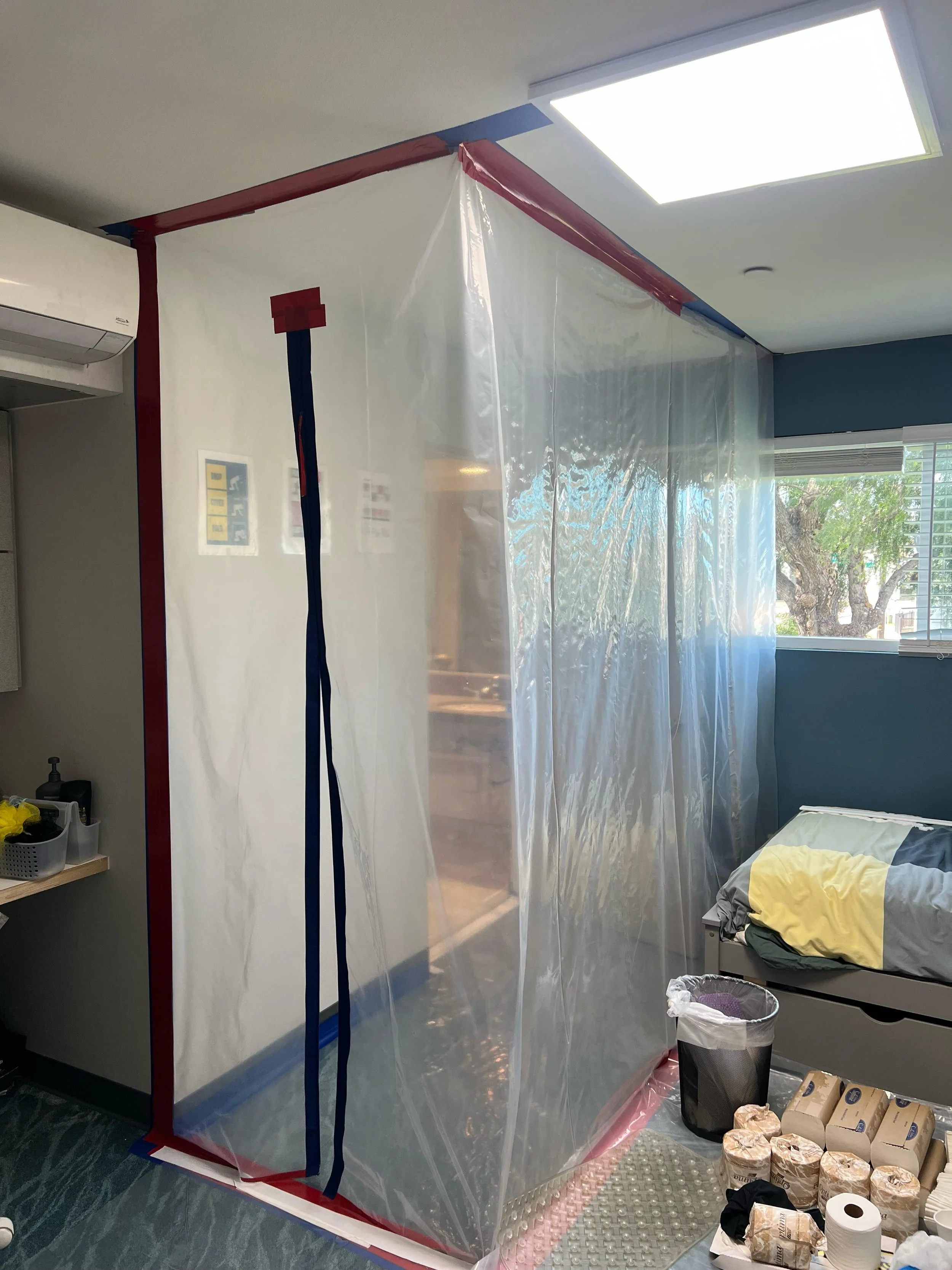 Room with a plastic partition, an air conditioning unit, a bed with yellow and gray bedding, a window with blinds, and various items like paper rolls, trash bin, and supplies.