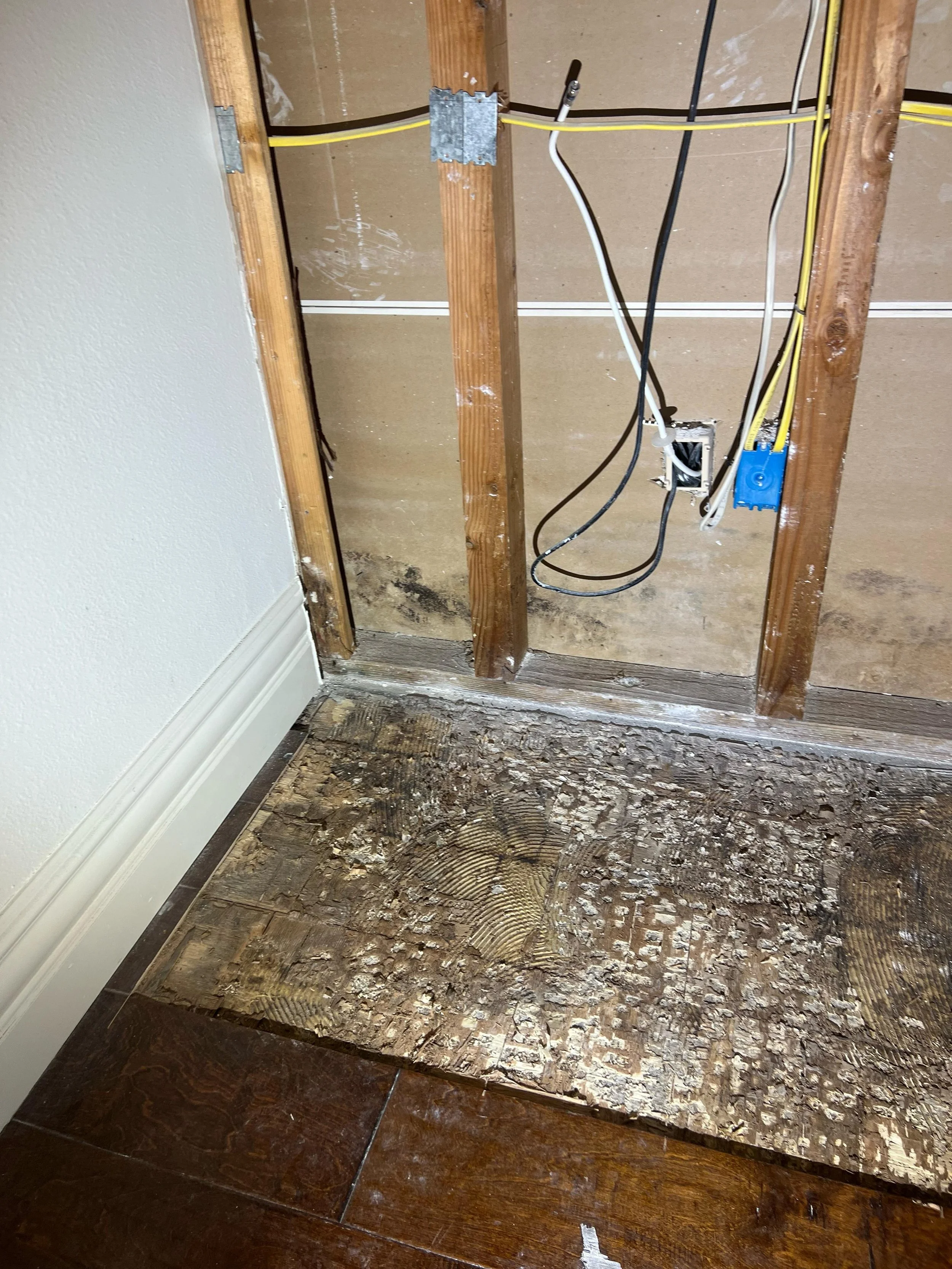 Partially constructed wall with exposed electrical wiring, electrical box, and wooden studs; the floor is partially removed, showing subflooring underneath and a white baseboard on the left side.