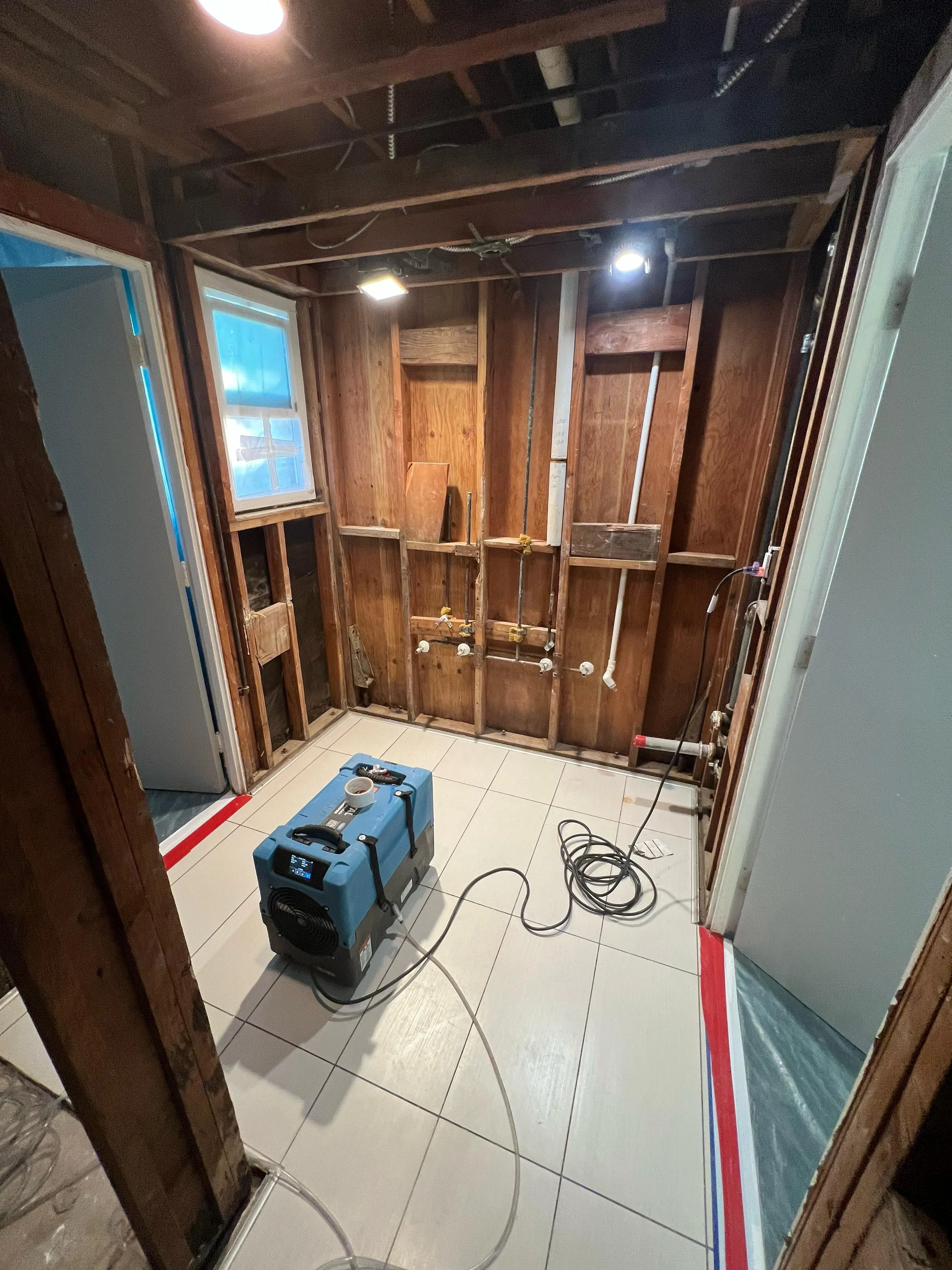 A partially renovated bathroom with exposed wooden wall studs and plumbing pipes. A window is present on the left side, and the floor has white tiles. A blue portable heater is on the floor, with extension cords and tools nearby. Red and blue painter