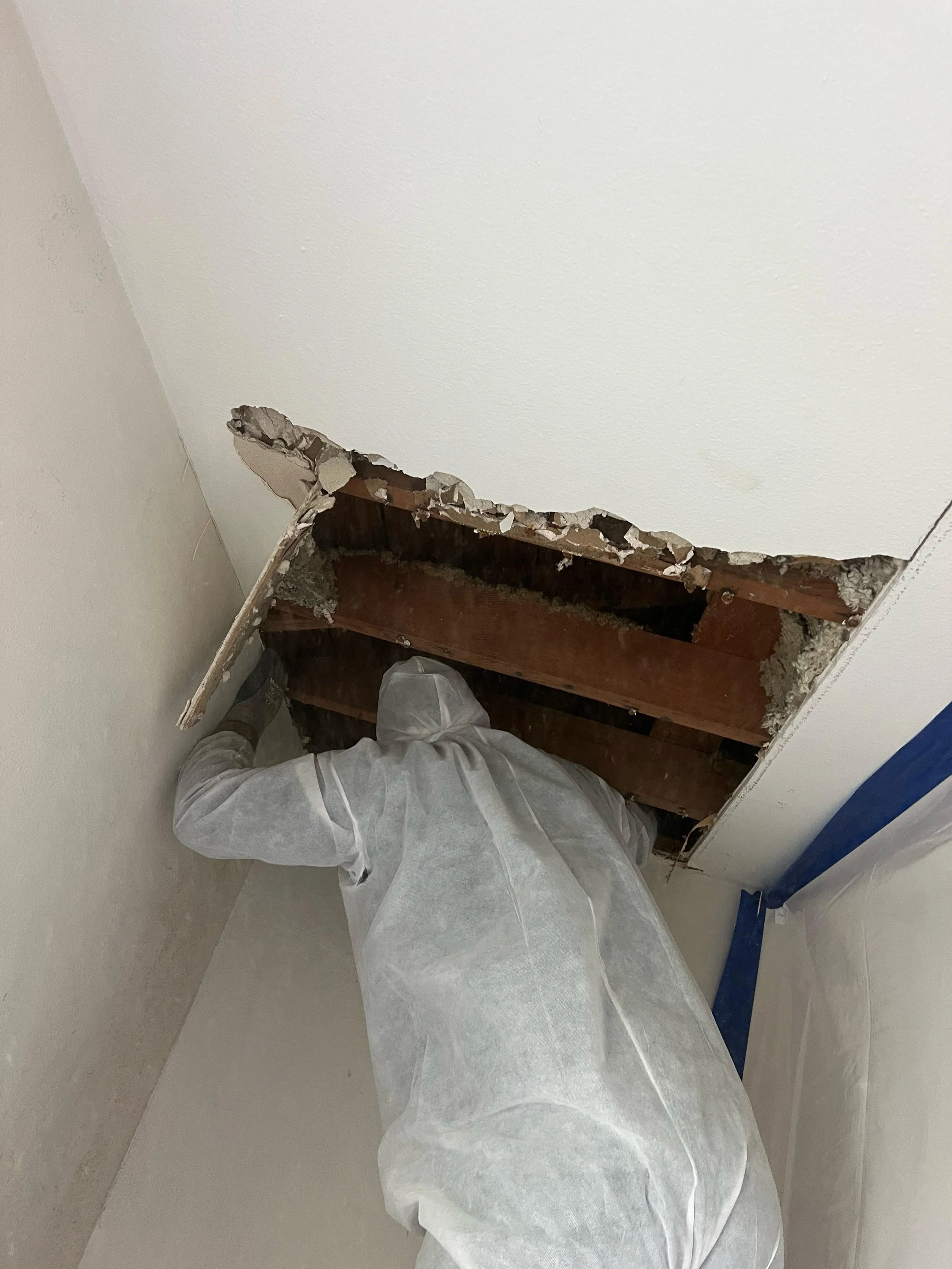 A construction worker in protective clothing is working on a drywall repair near the floor. The wall has a large hole exposing wooden studs and pipes, with debris and dust around the area.