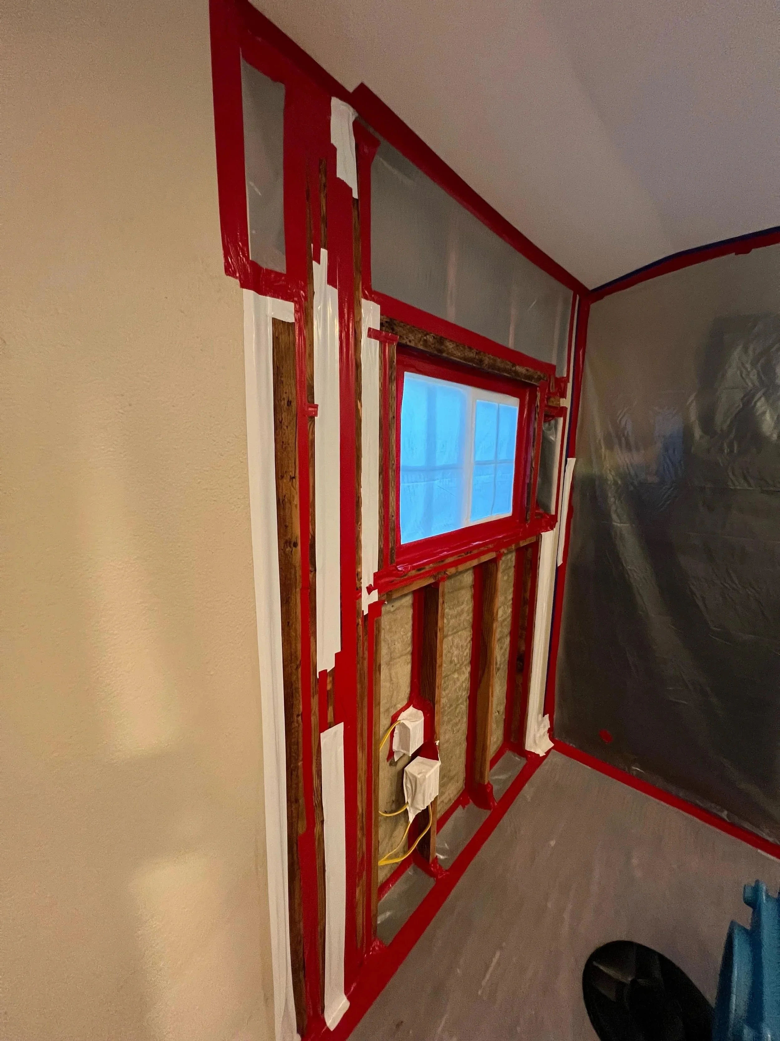 Interior view of a wall under renovation, with exposed studs, insulation, electrical wiring, and plastic sheeting, partially covered by painter's tape.
