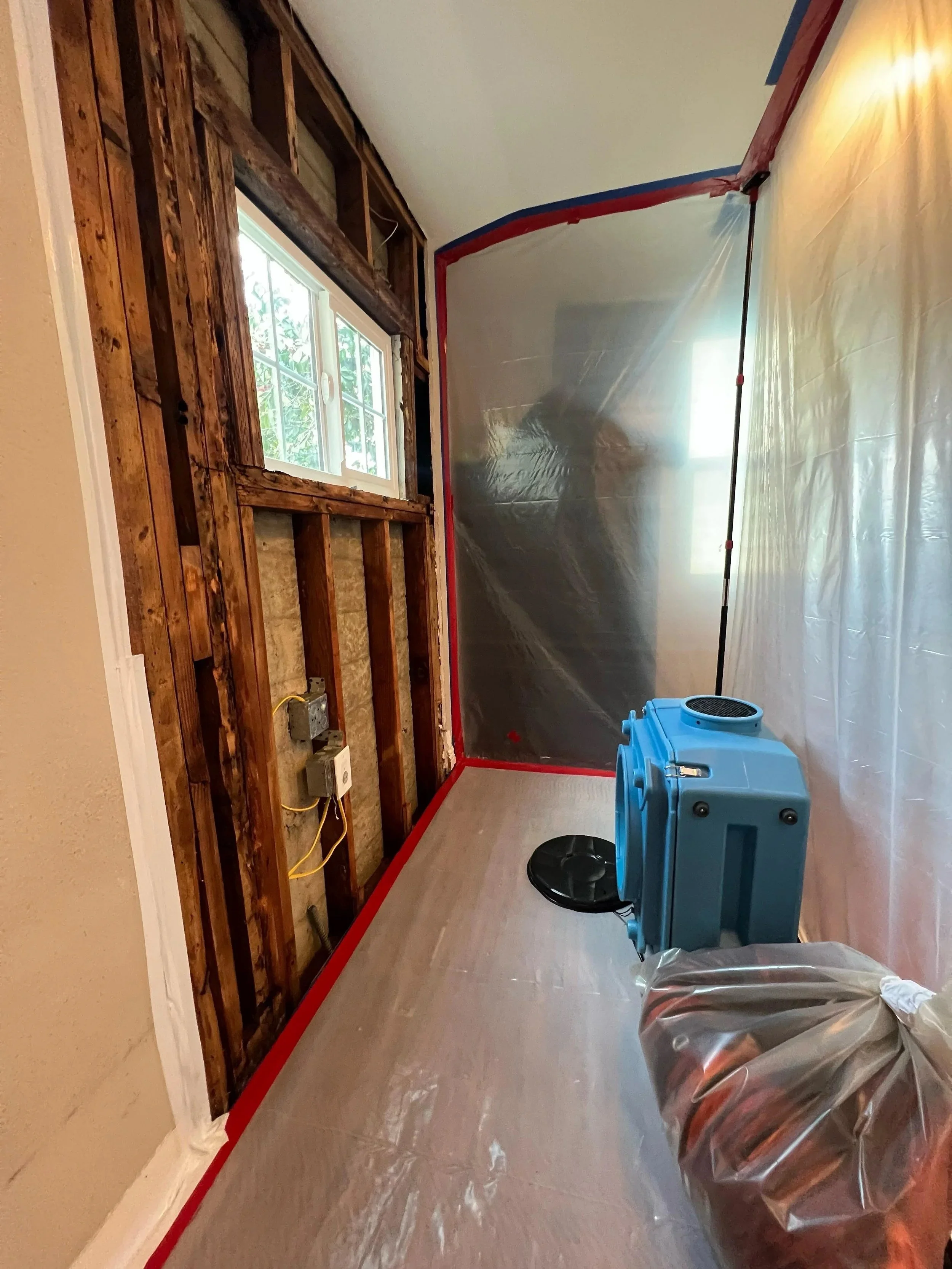A room under renovation with exposed wall studs, partial window, plastic sheet on part of the wall, a portable air purifier, and a covered object in the corner.