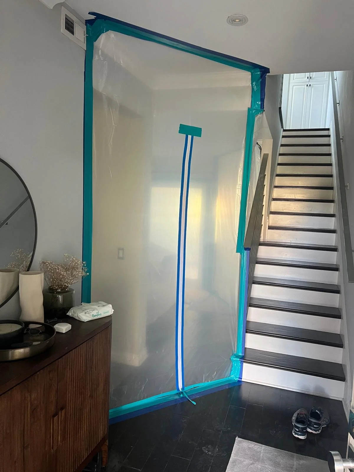 Clear plastic sheet with blue painter's tape around edges, hanging as a temporary barrier next to a staircase in a home interior.