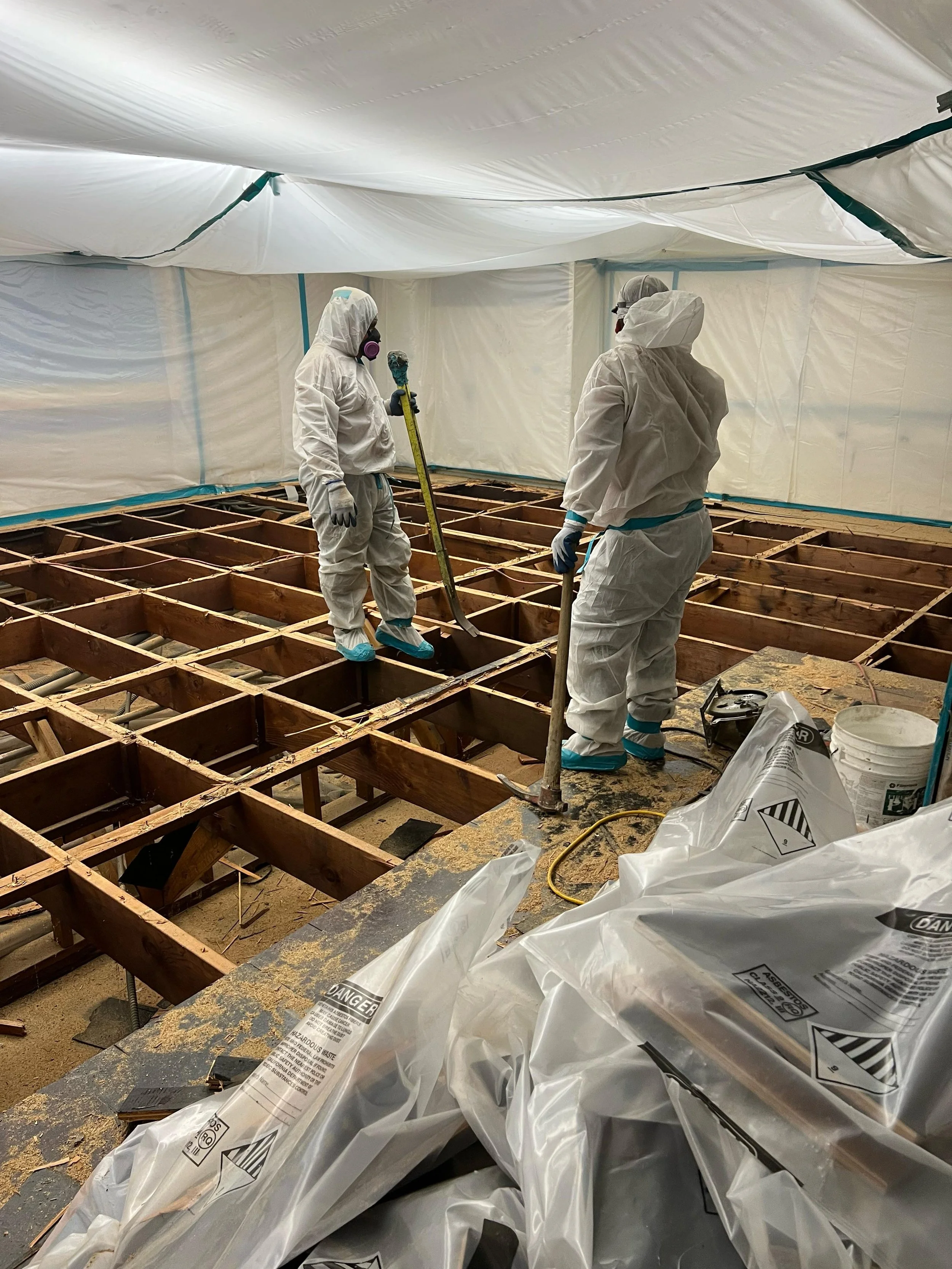 Two workers in protective suits and masks working on a construction floor with exposed wood framing, plastic sheeting on the ceiling and walls, and construction materials in the foreground.