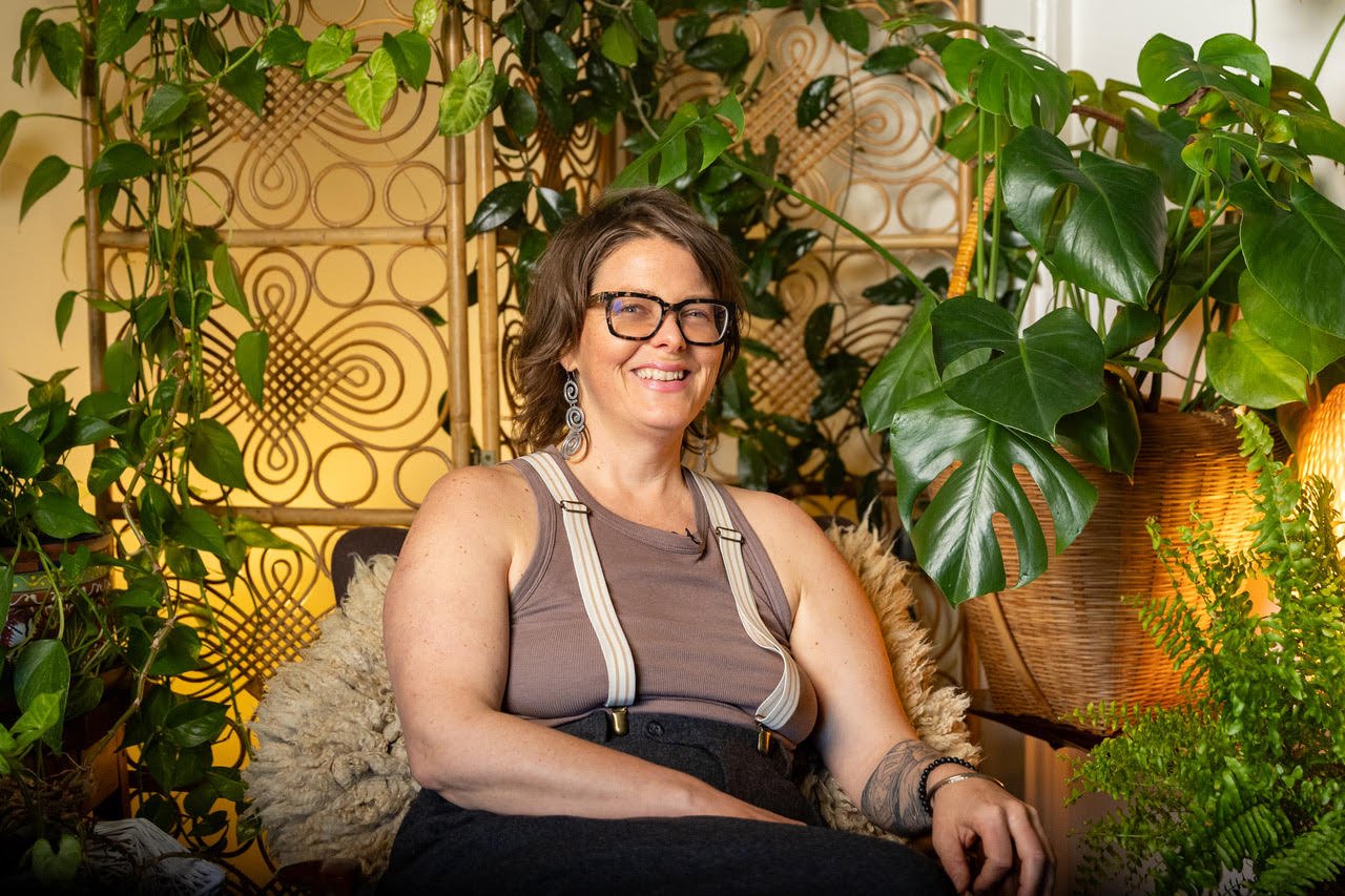 A woman with glasses and earrings sitting on a chair, surrounded by green plants in a cozy indoor space.