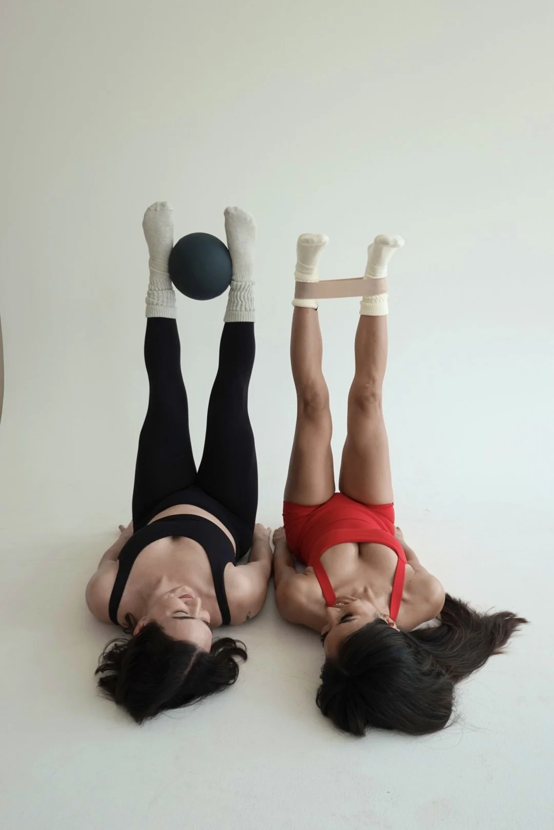 Two women lying on their backs with legs raised, using resistance bands and a ball for exercise, against a plain wall background.
