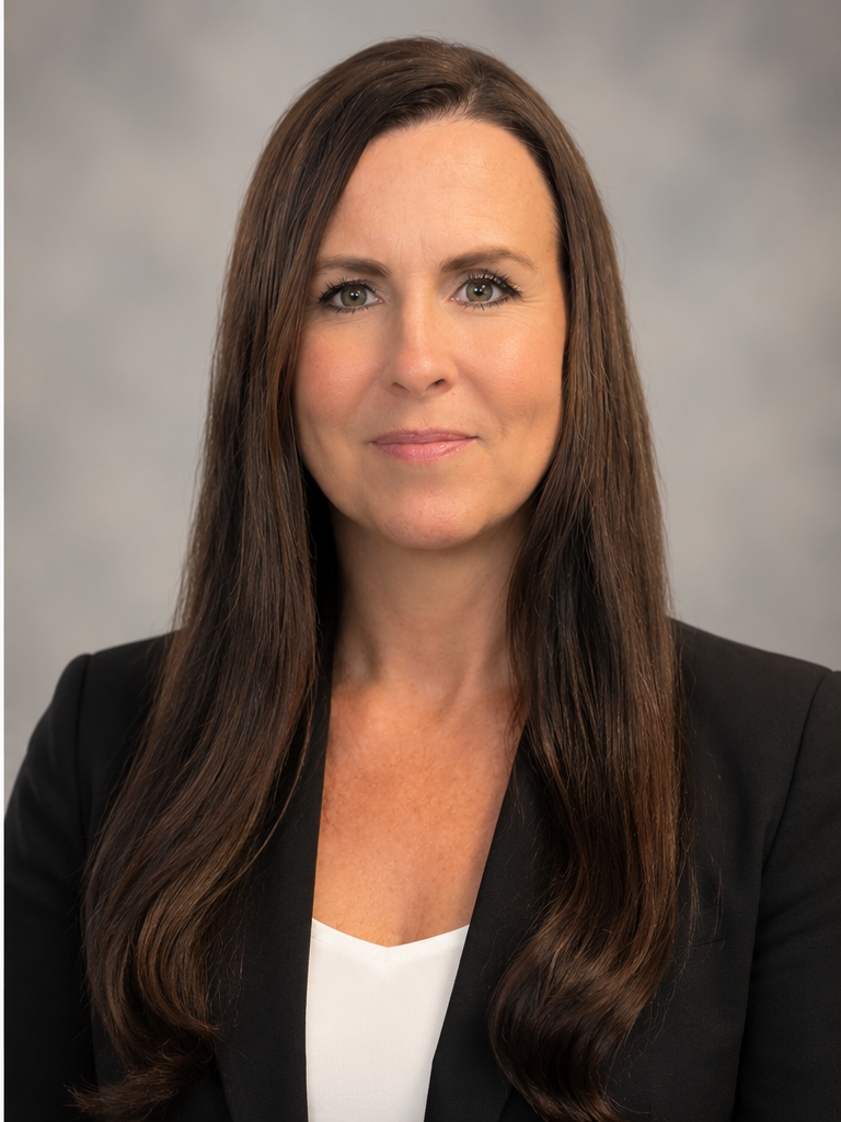A woman with long brown hair and light skin, wearing a black blazer over a white top, looking into the camera with a neutral expression, against a plain gray background.