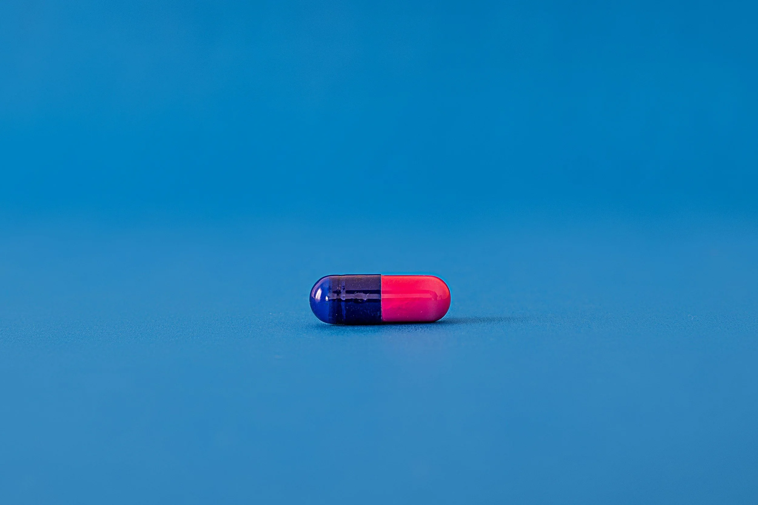 A capsule pill with half blue and half pink coloring resting on a plain blue background.