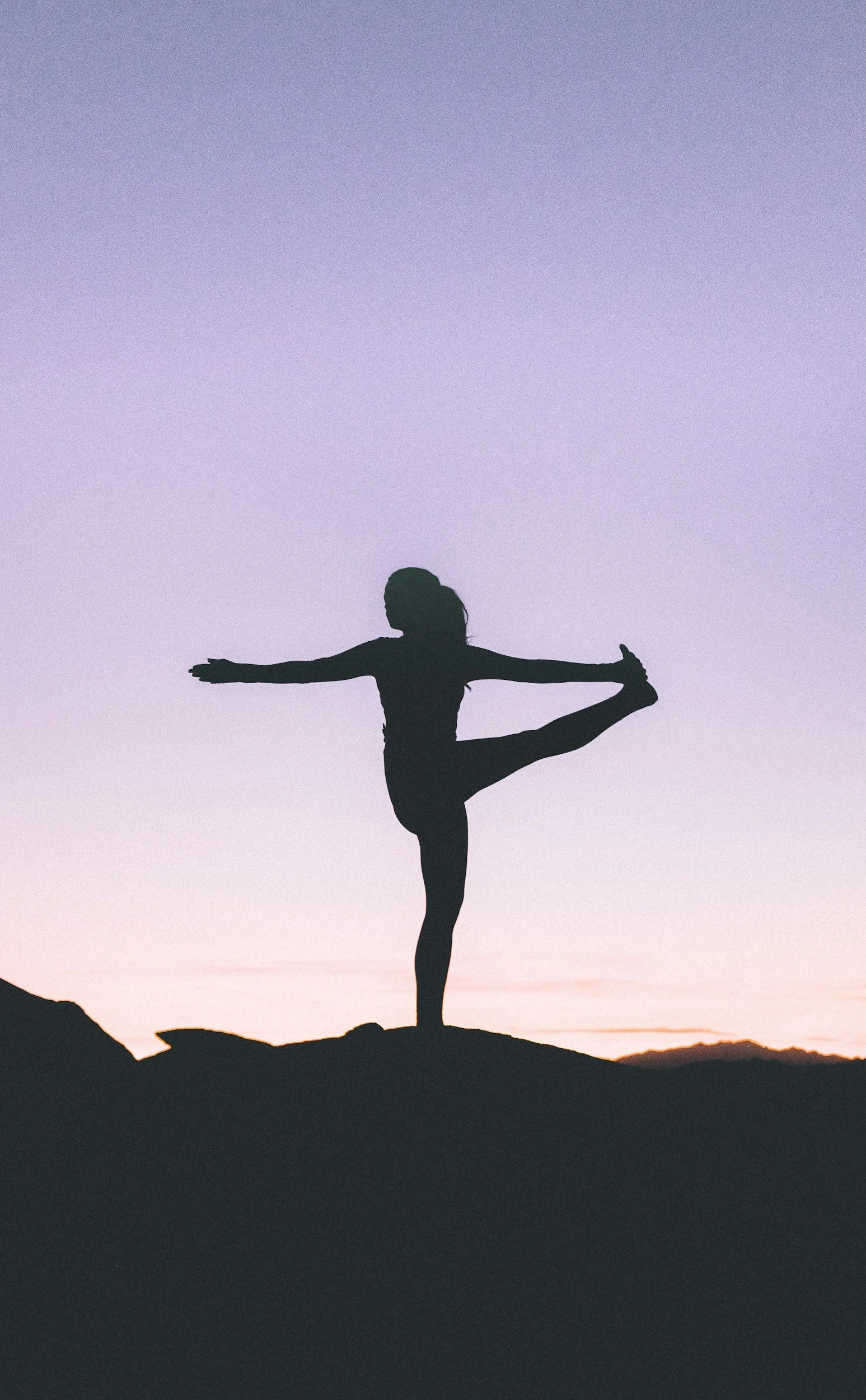Silhouette of a person performing a yoga pose, standing on one leg, with arms extended and the other leg bent behind, against a pastel-colored sunset or sunrise sky with distant mountains.