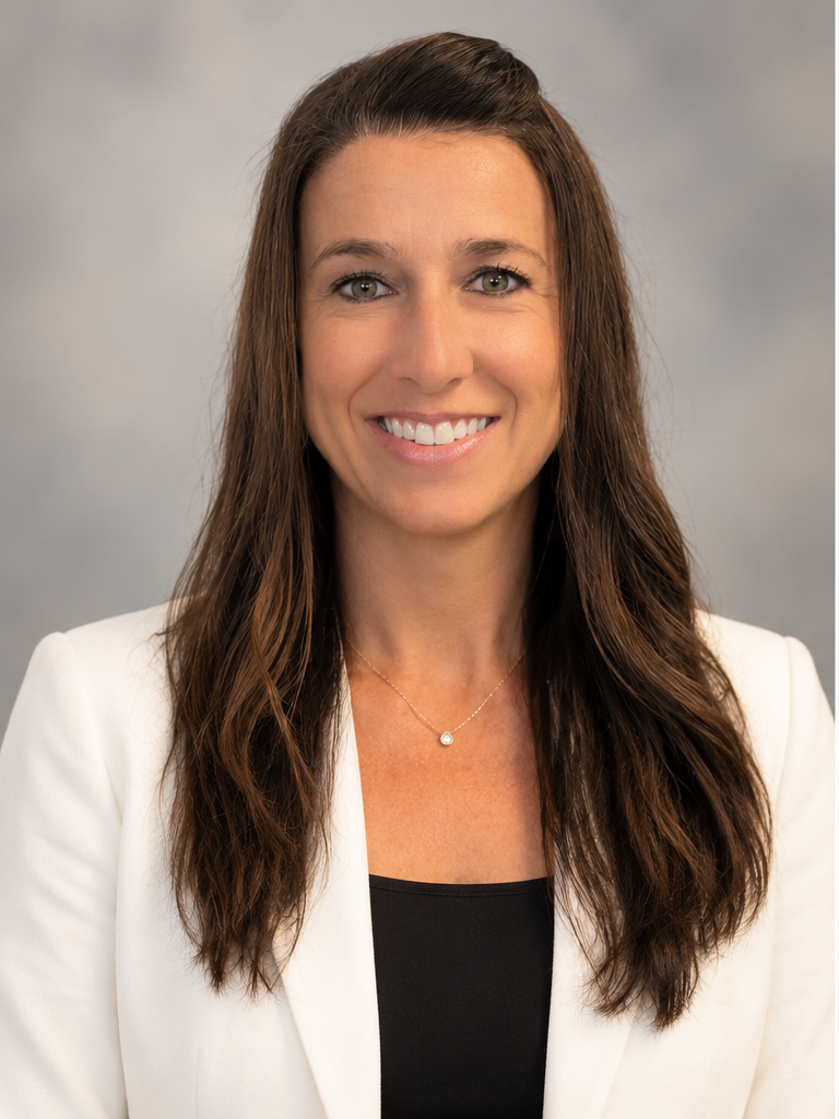 A woman with long brown hair, wearing a white blazer and a black top, smiling for a professional portrait.