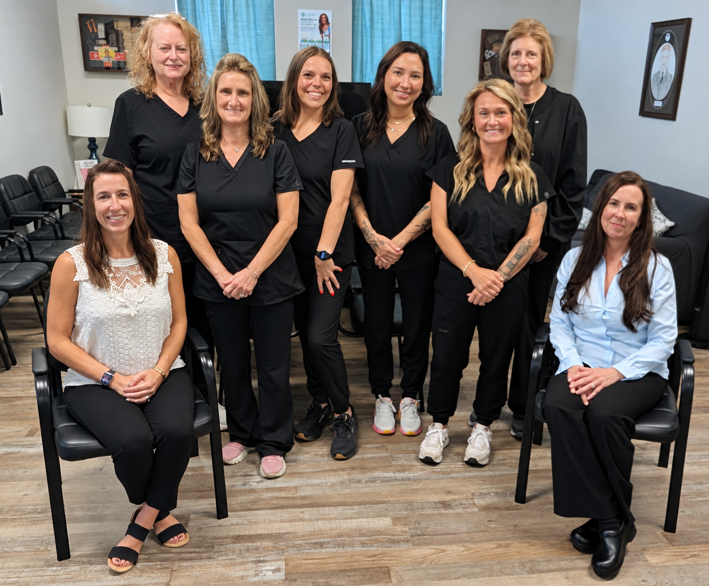 Group of ten women, some wearing black scrubs, posed in a room with chairs, framed pictures, and a blue curtain in the background.