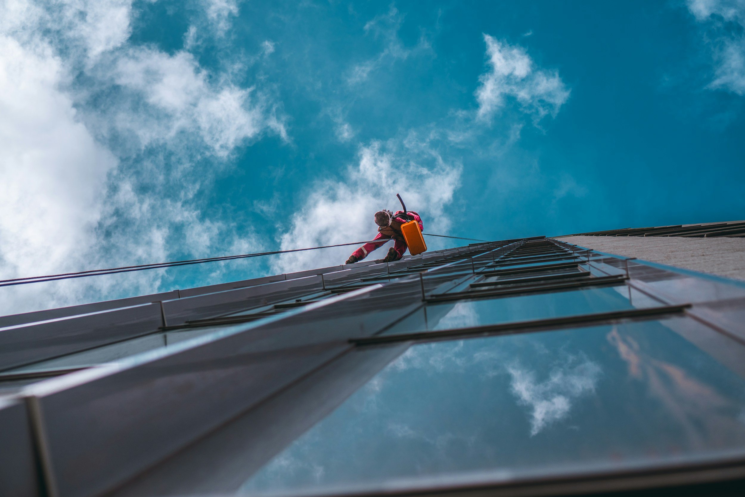 A person cleaning the exterior glass of a tall building with a harness and cleaning equipment, against a blue sky with clouds.