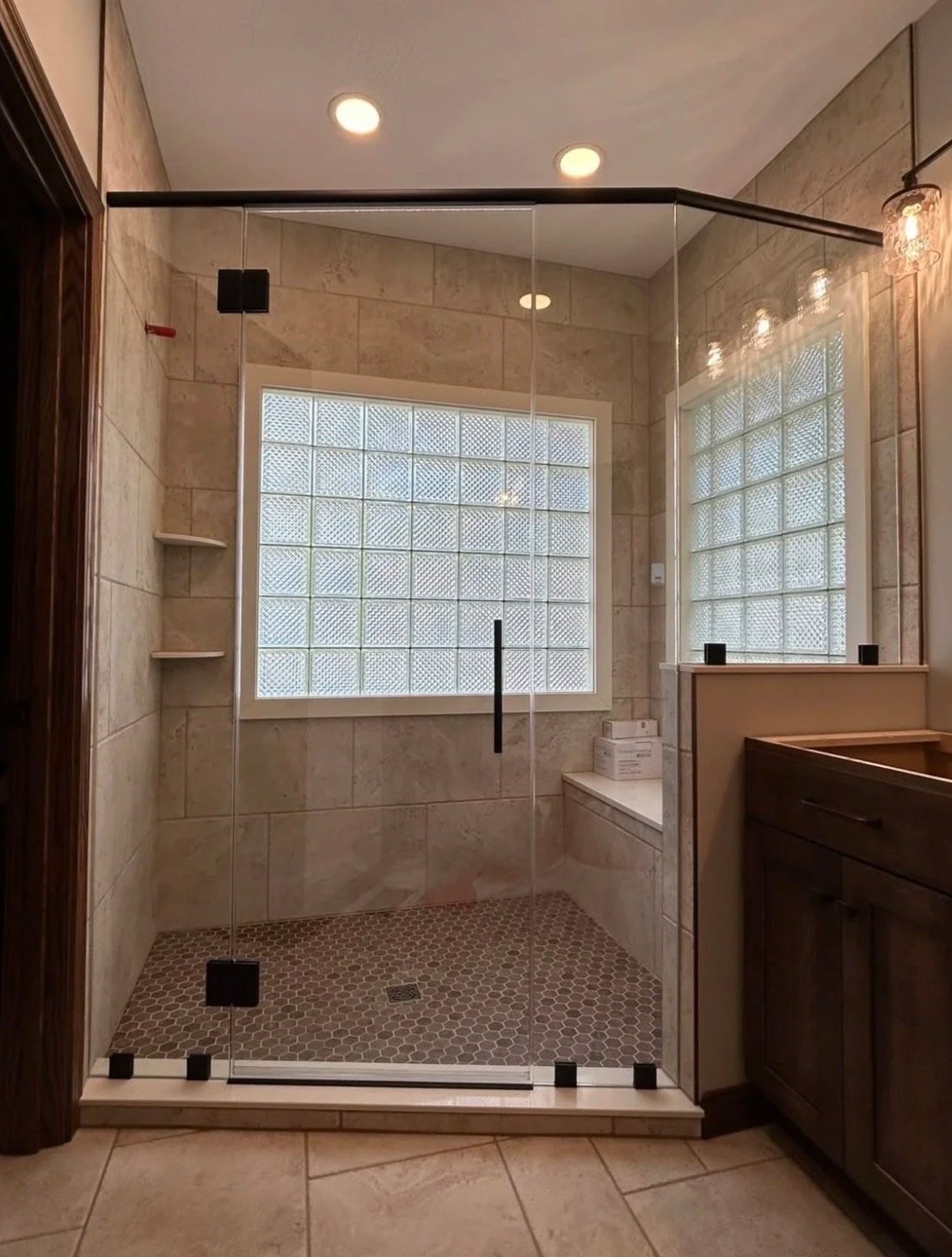 Bathroom with glass shower enclosure, beige tiled walls, glass block windows, small hexagonal floor tiles, and a wooden vanity with a mirror.