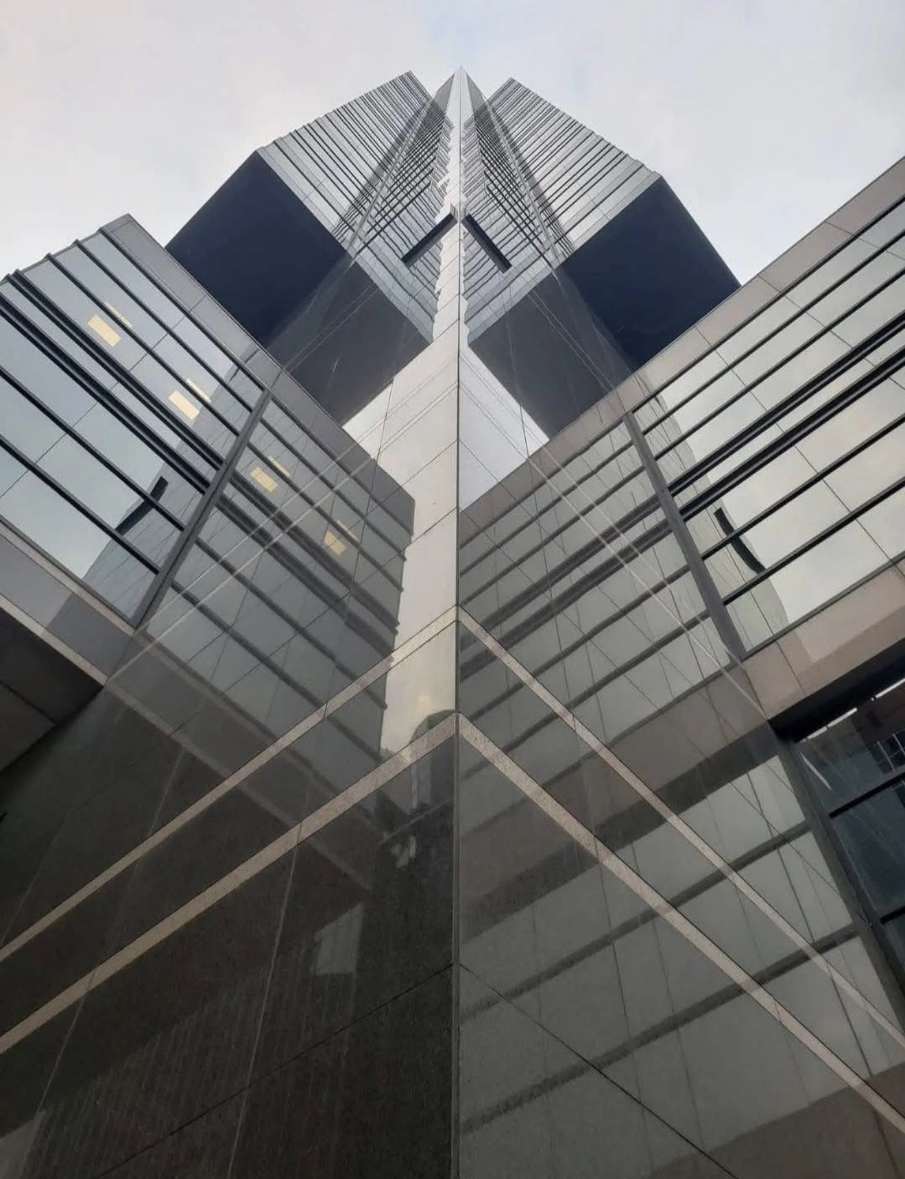 Looking up at a modern glass skyscraper with reflective windows, symmetrical architecture, and a central mirror-like surface that creates a mirror image.