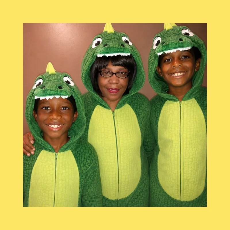 Three people dressed in matching green dinosaur costumes with hoods that have dinosaur faces and teeth, standing together indoors, smiling at the camera.