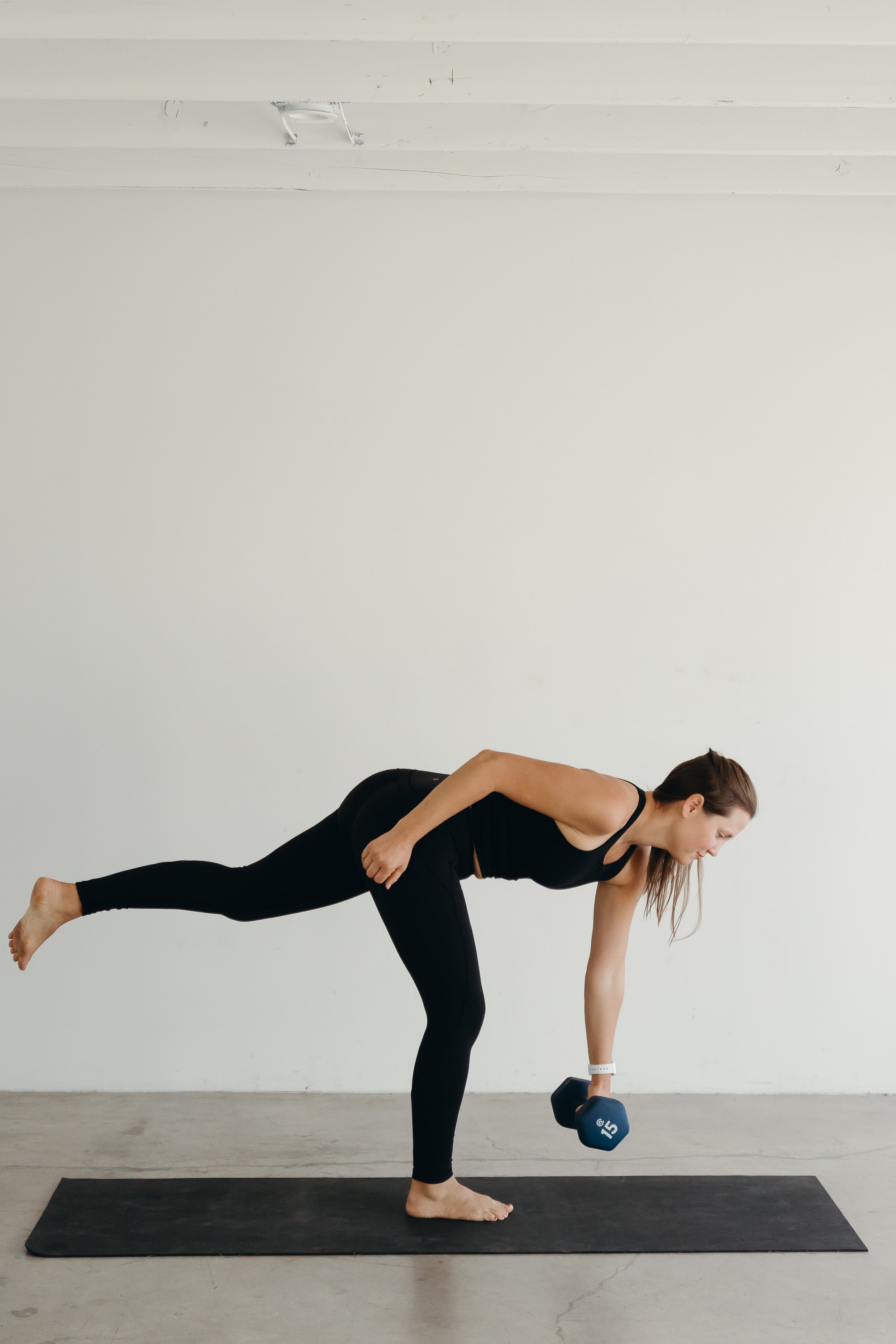 Woman in black workout clothes performing a single-leg deadlift with a dumbbell in a bright, minimal studio with white walls and a black exercise mat.