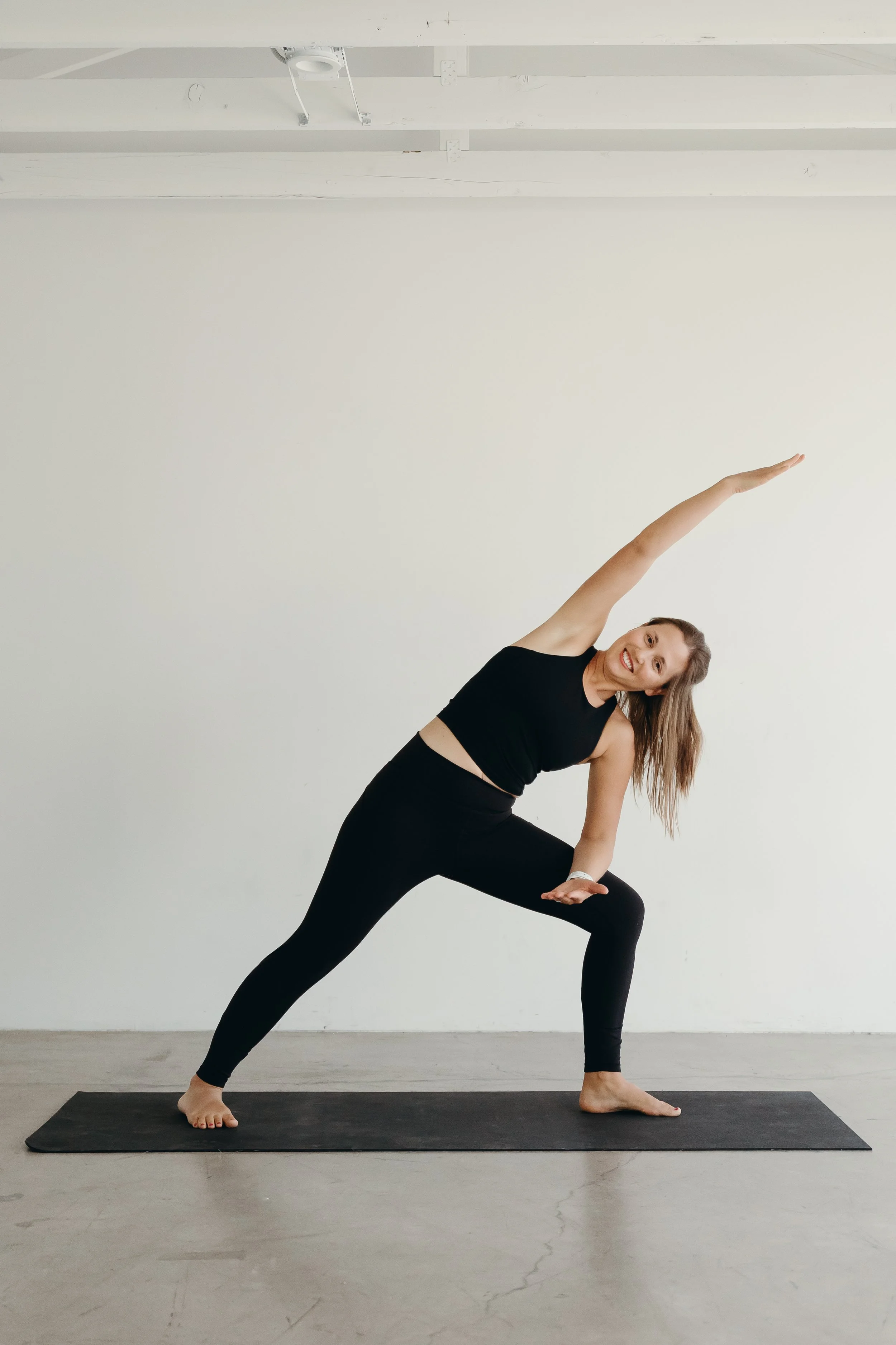 A woman practicing yoga indoors on a black yoga mat, standing in a side lunge position with her right leg bent and left leg extended back. She is wearing black athletic wear and smiling with her arms raised, one arm reaching up and the other extended downward.