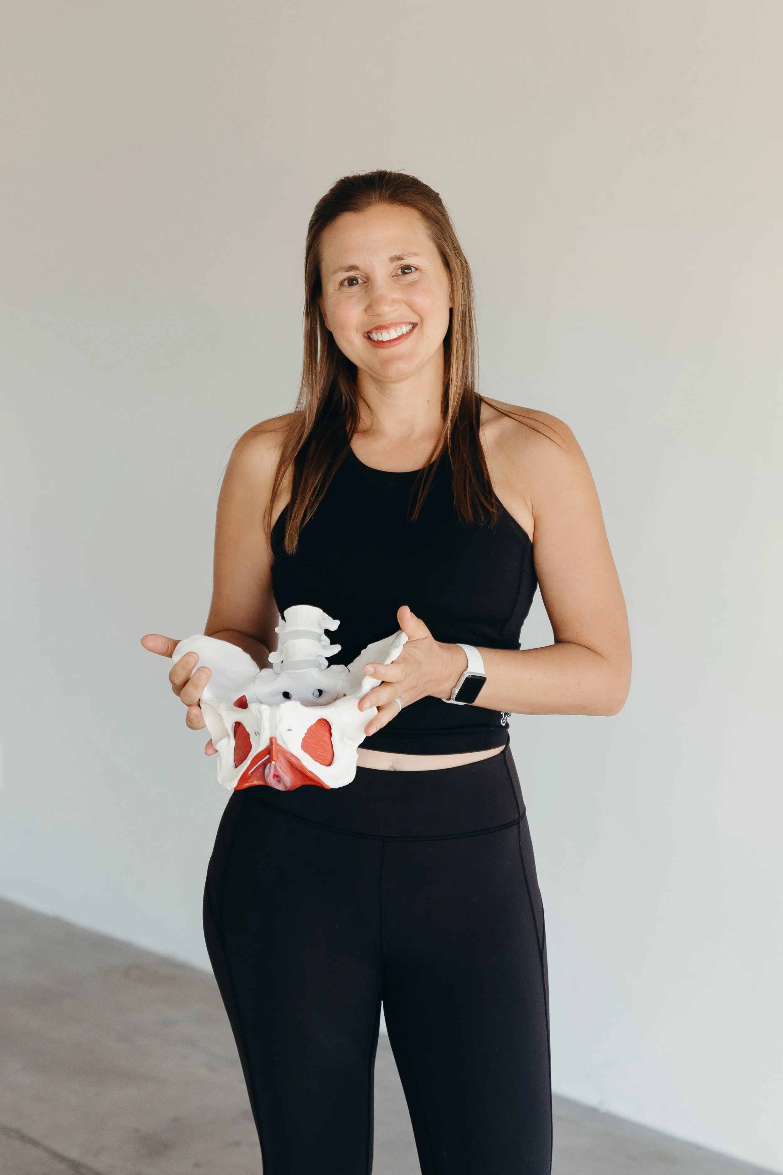 A woman holding a model of a human pelvis and spine, standing in front of a plain wall, smiling and wearing a black workout outfit.