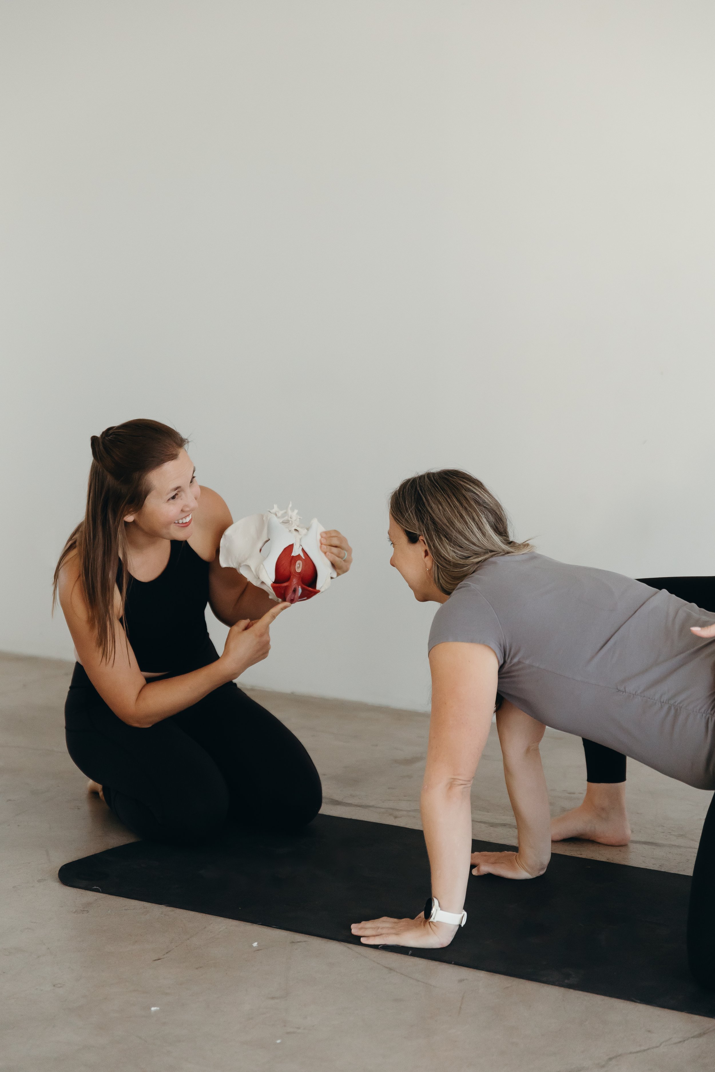 A woman kneeling on a yoga mat holding a model of the human pelvis and discussing it with another woman performing a yoga pose.