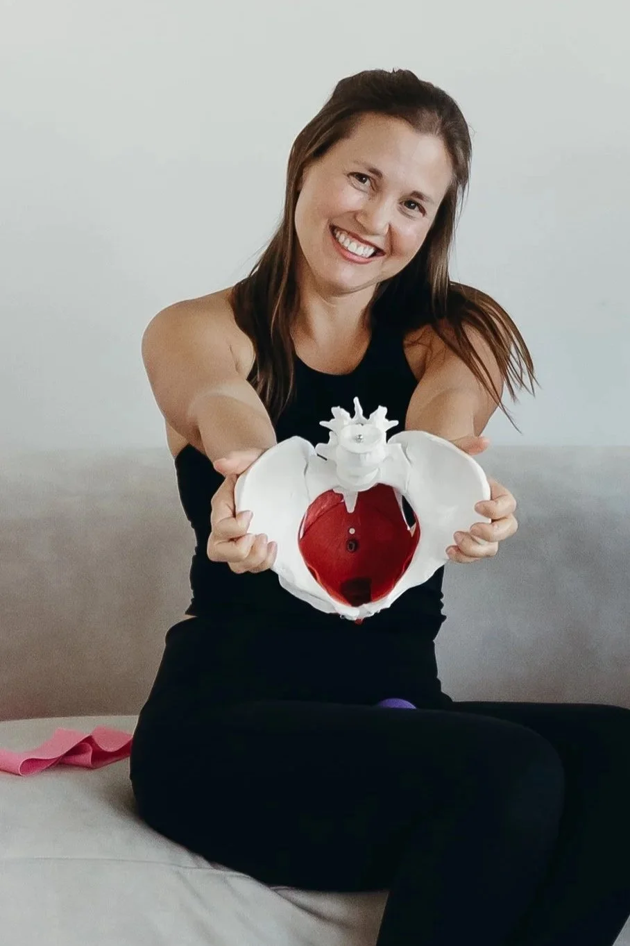 A smiling woman sitting on a bed, holding a model of a human heart facing the camera, wearing a black tank top and black leggings.