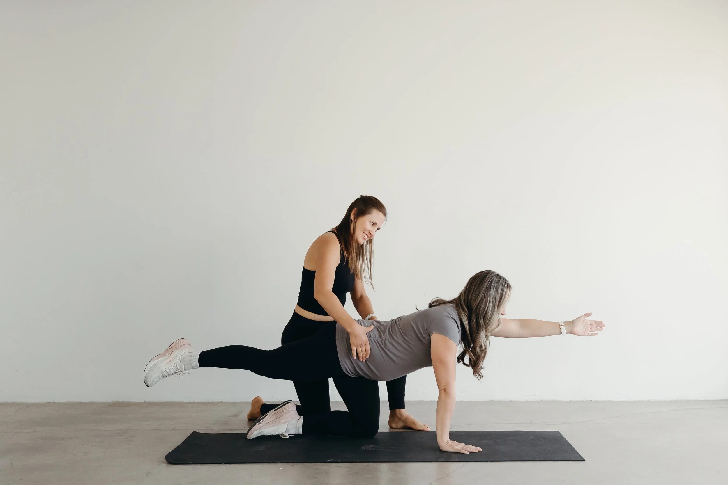 Two women practicing yoga indoors on a black mat. The woman in the front is on all fours with her right arm extended forward and her body parallel to the floor. The woman behind is standing, supporting the first woman's waist with her hands, and smil