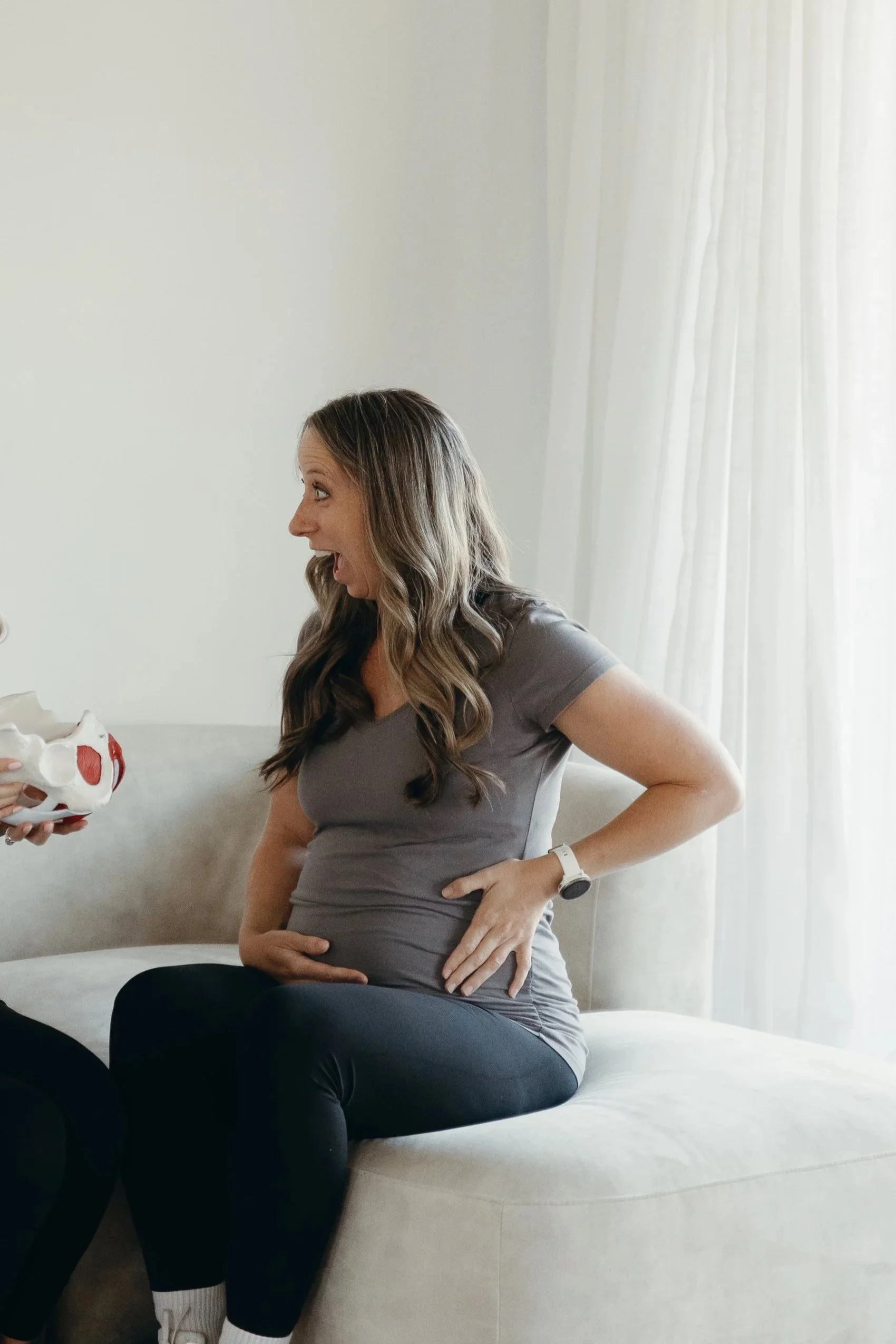 A woman with long brown hair, wearing a gray shirt and black pants, sitting on a bed with her hand on her pregnant belly, talking to someone holding a model of a pelvis.