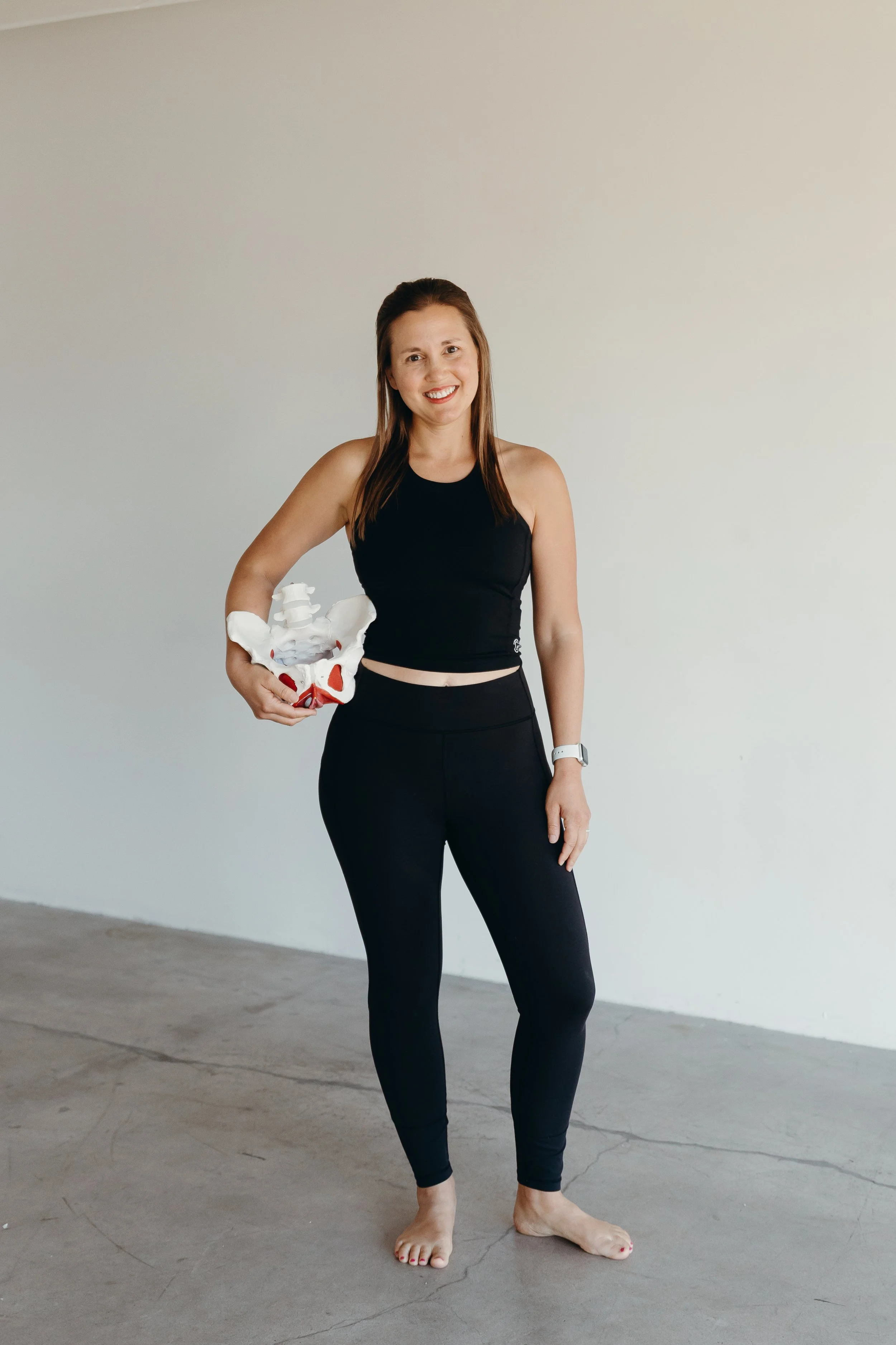 A woman with brown hair, wearing black workout clothes, barefoot, standing in a room with plain white walls. She is smiling and holding a model of a human pelvis in her hand.