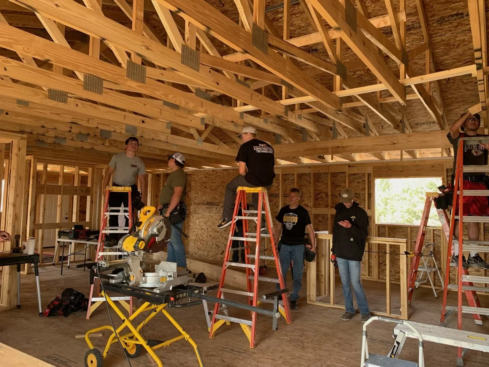 Construction workers building the wooden framework of a house interior, using ladders, power tools, and other tools inside a partially constructed home.
