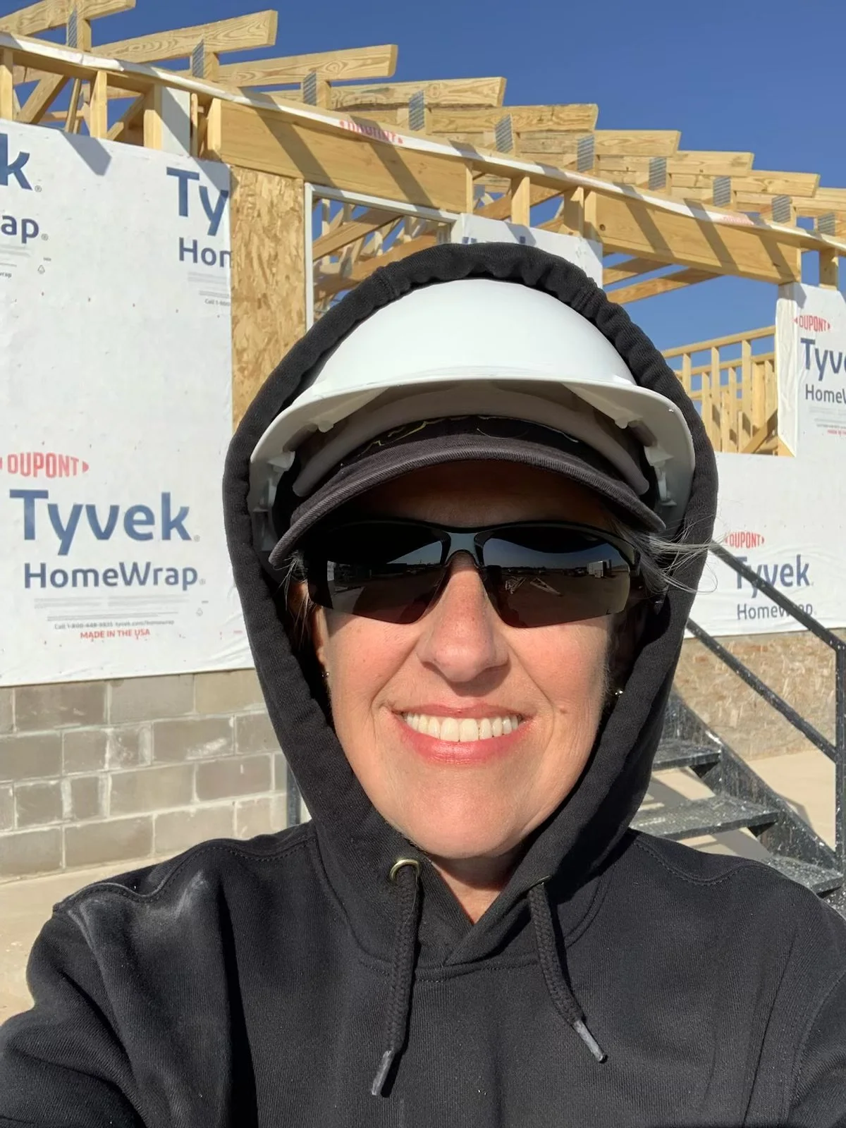 A woman smiling at the camera wearing sunglasses, a hoodie with the hood up, and a white helmet at a building construction site with wooden framing and Tyvek HomeWrap.