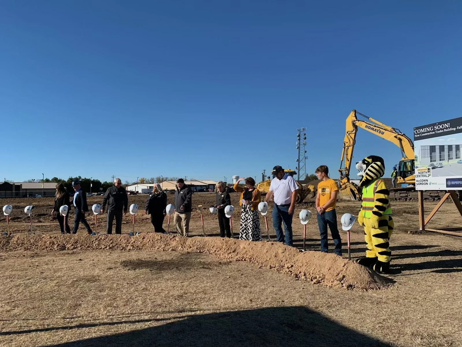 People participating in a ground-breaking ceremony at a construction site, with a person in a tiger mascot costume and a large construction digger in the background.