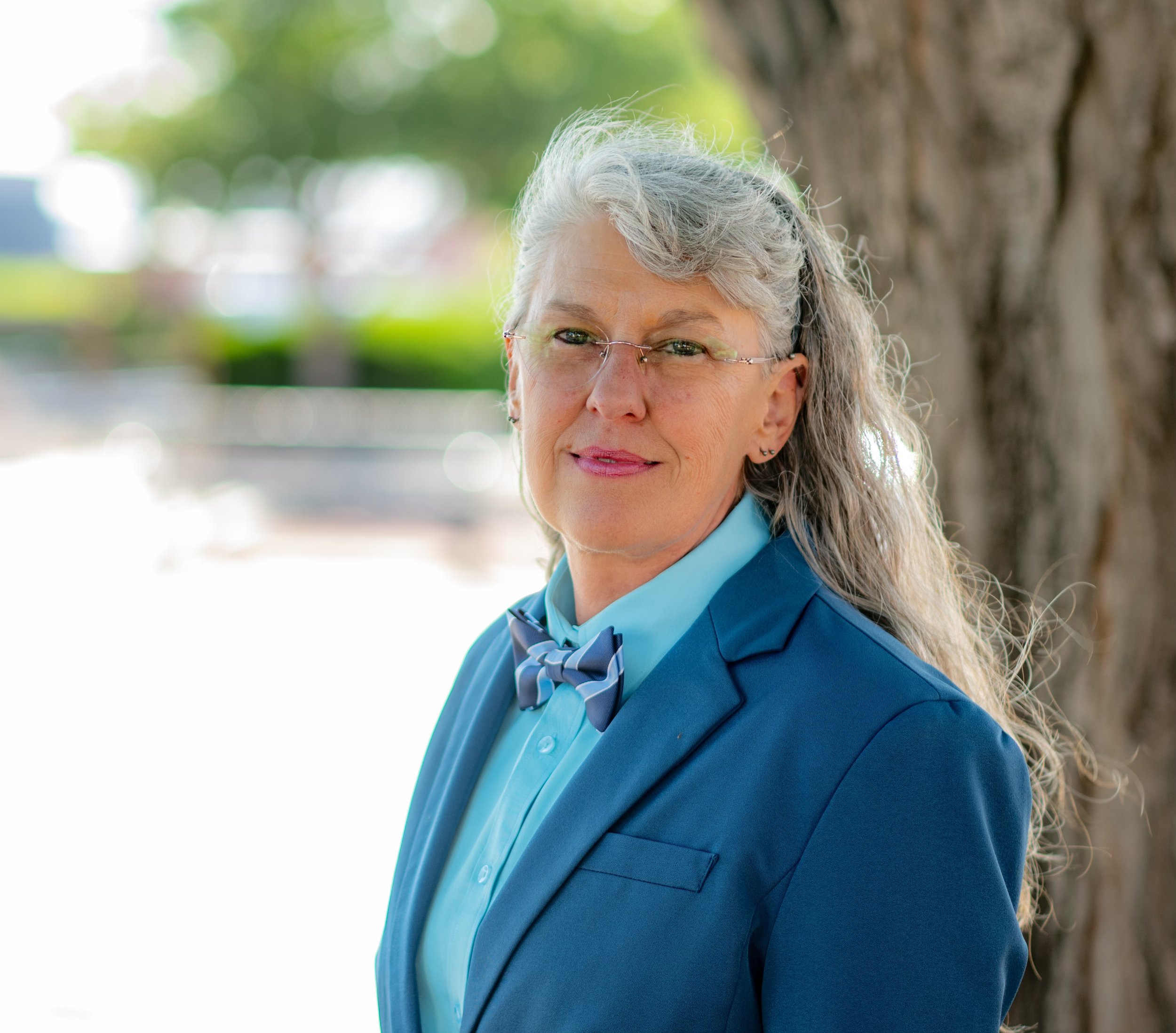 Close-up of an older woman with gray hair, glasses, and a blue blazer outdoors, smiling softly with a tree in the background.