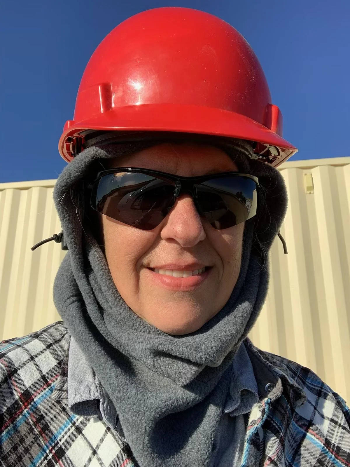 Person wearing a red safety helmet, black sunglasses, gray hoodie, and plaid shirt standing outdoors with a blue sky and beige building in the background.