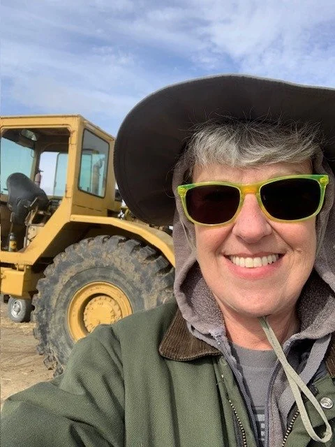 A smiling woman wearing a wide-brimmed hat, sunglasses, and a jacket, standing outdoors next to a yellow construction vehicle, possibly a bulldozer, under a partly cloudy sky.