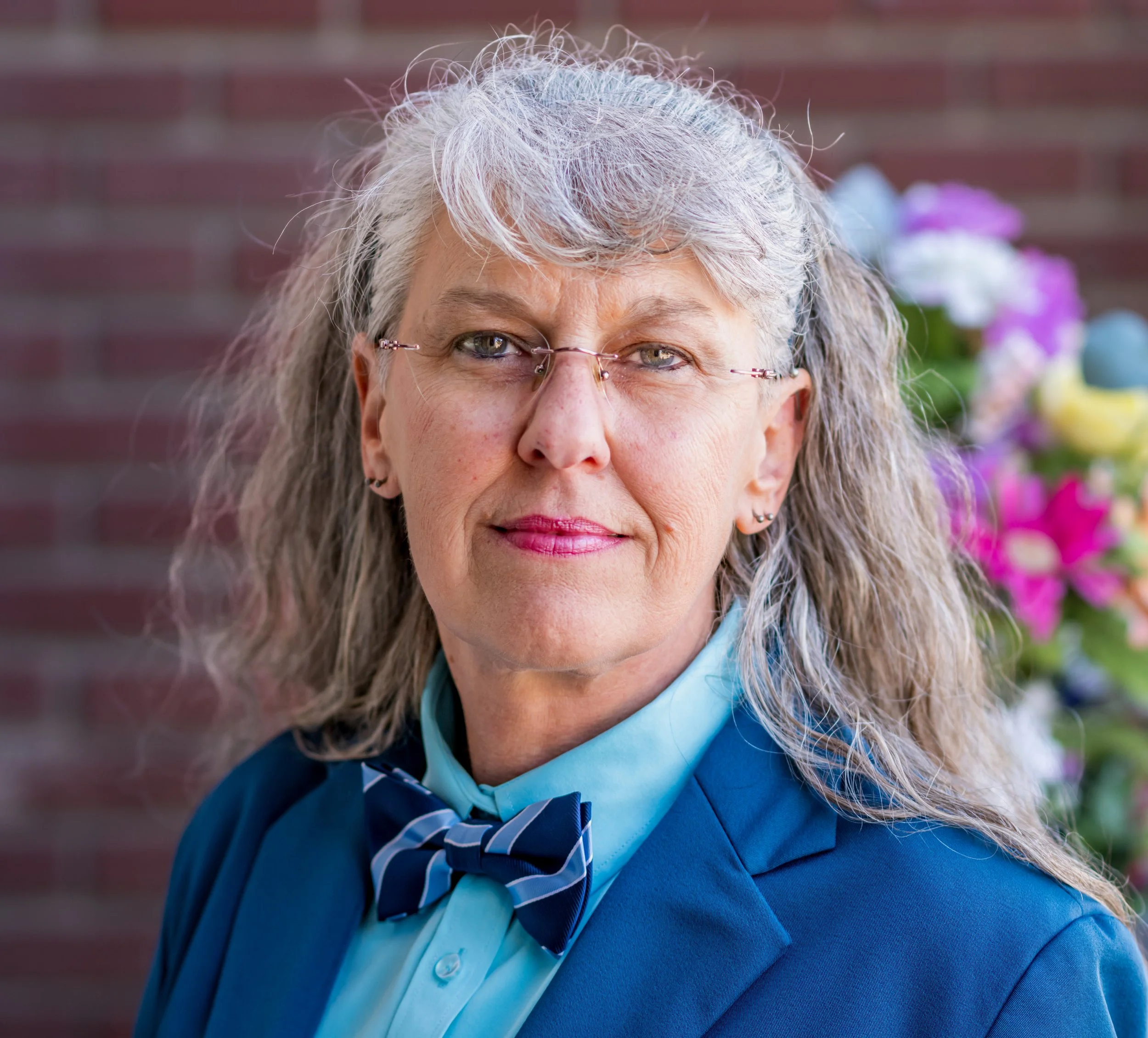 Close-up of an older woman with gray, curly hair and glasses, wearing a blue jacket and a blue scarf with a white shirt, standing outdoors with a blurred brick wall and flowers in the background.